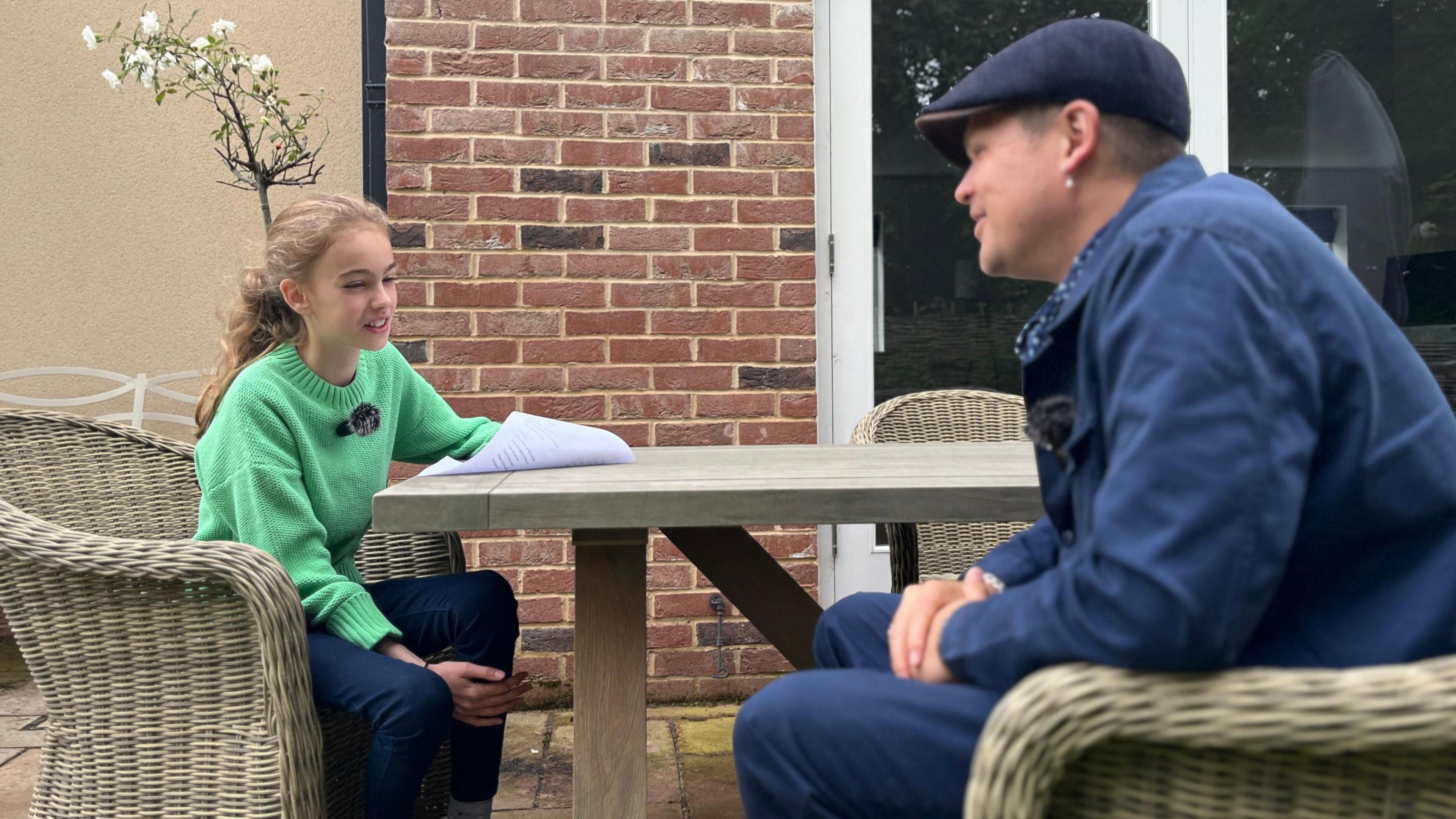 Anna, an 11 year old girl, wears a green jumper and sits at a garden table with poet Luke Wright