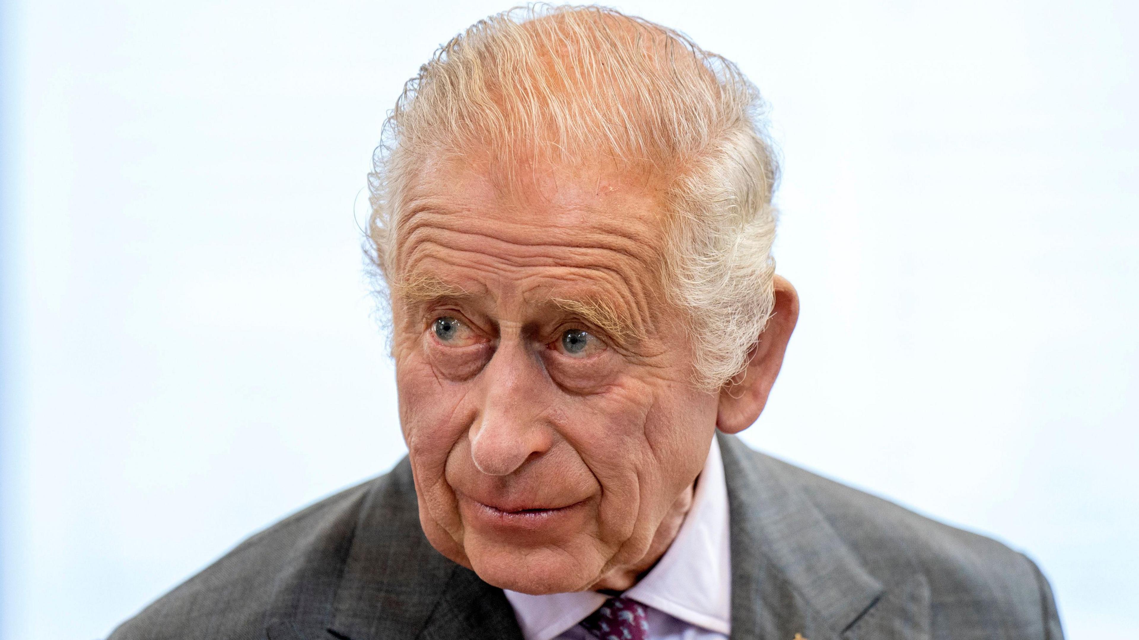 King Charles III looks off camera. He is wearing a shirt and tie and a dark coloured jacket over the top.