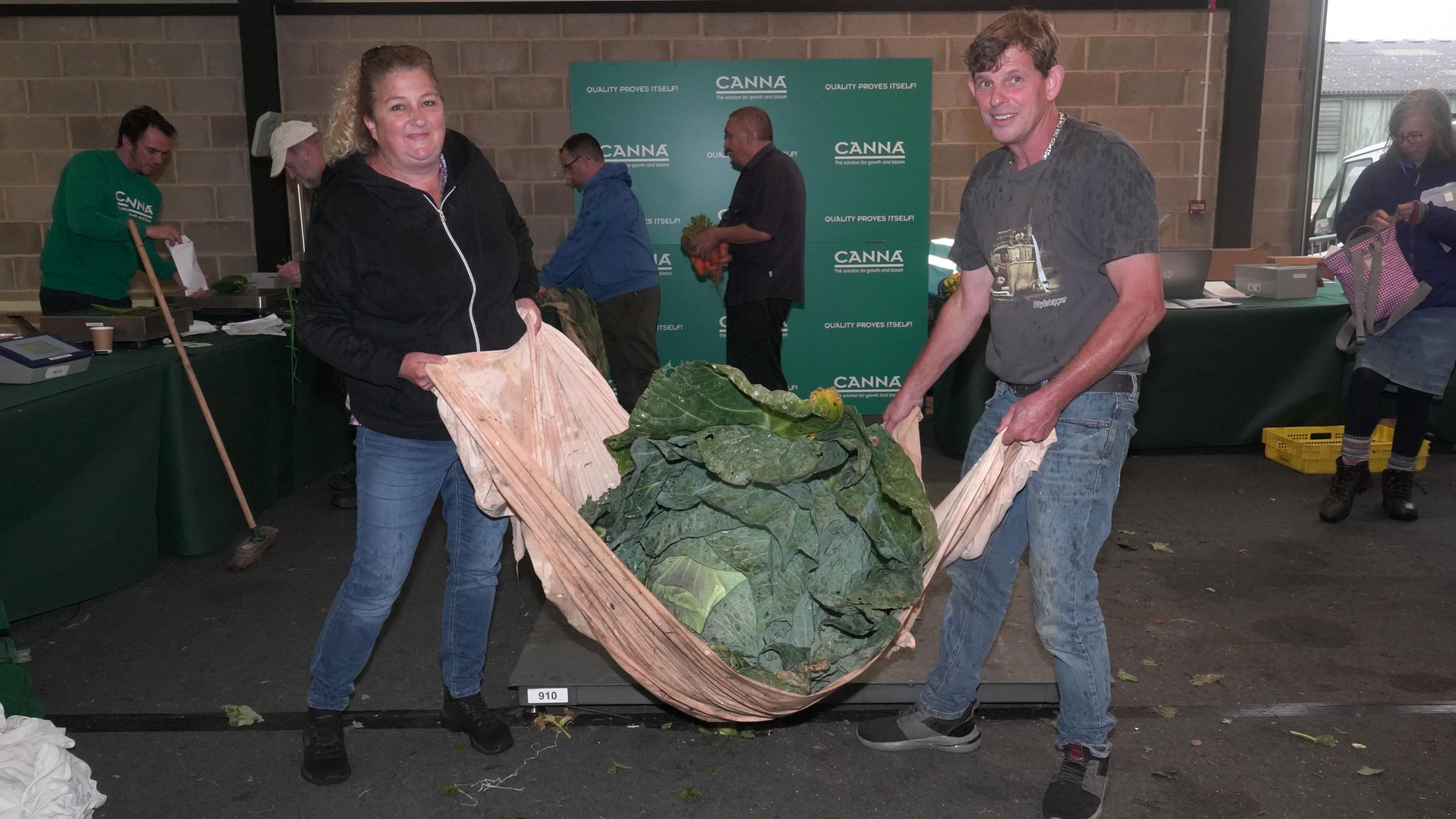 A woman and a man holding an enormous cabbage, lifting it from either side using a large brown cloth.