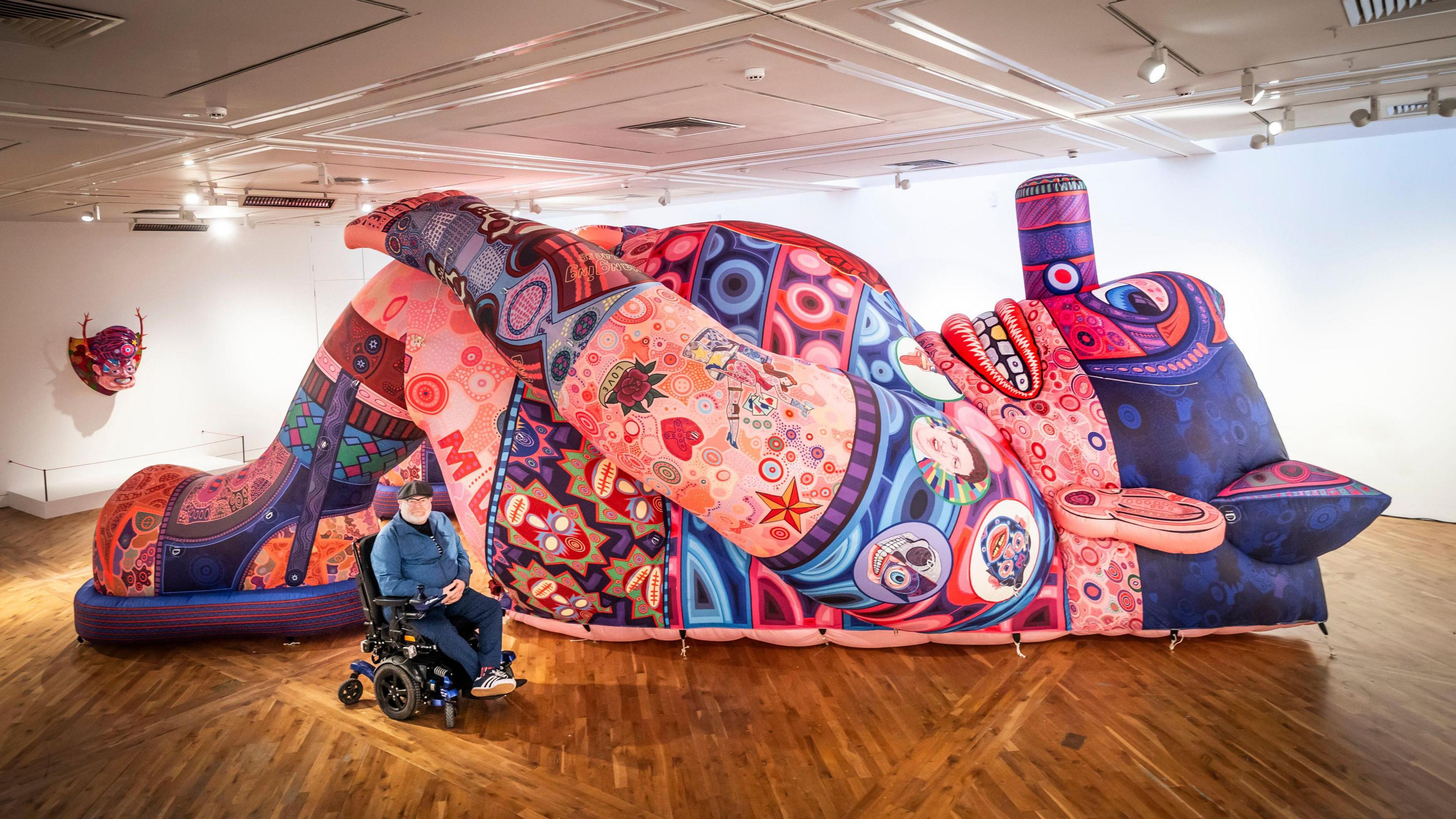 A man in a wheelchair poses with one of his large inflatable artworks, which stands as tall as the ceiling in an art gallery.