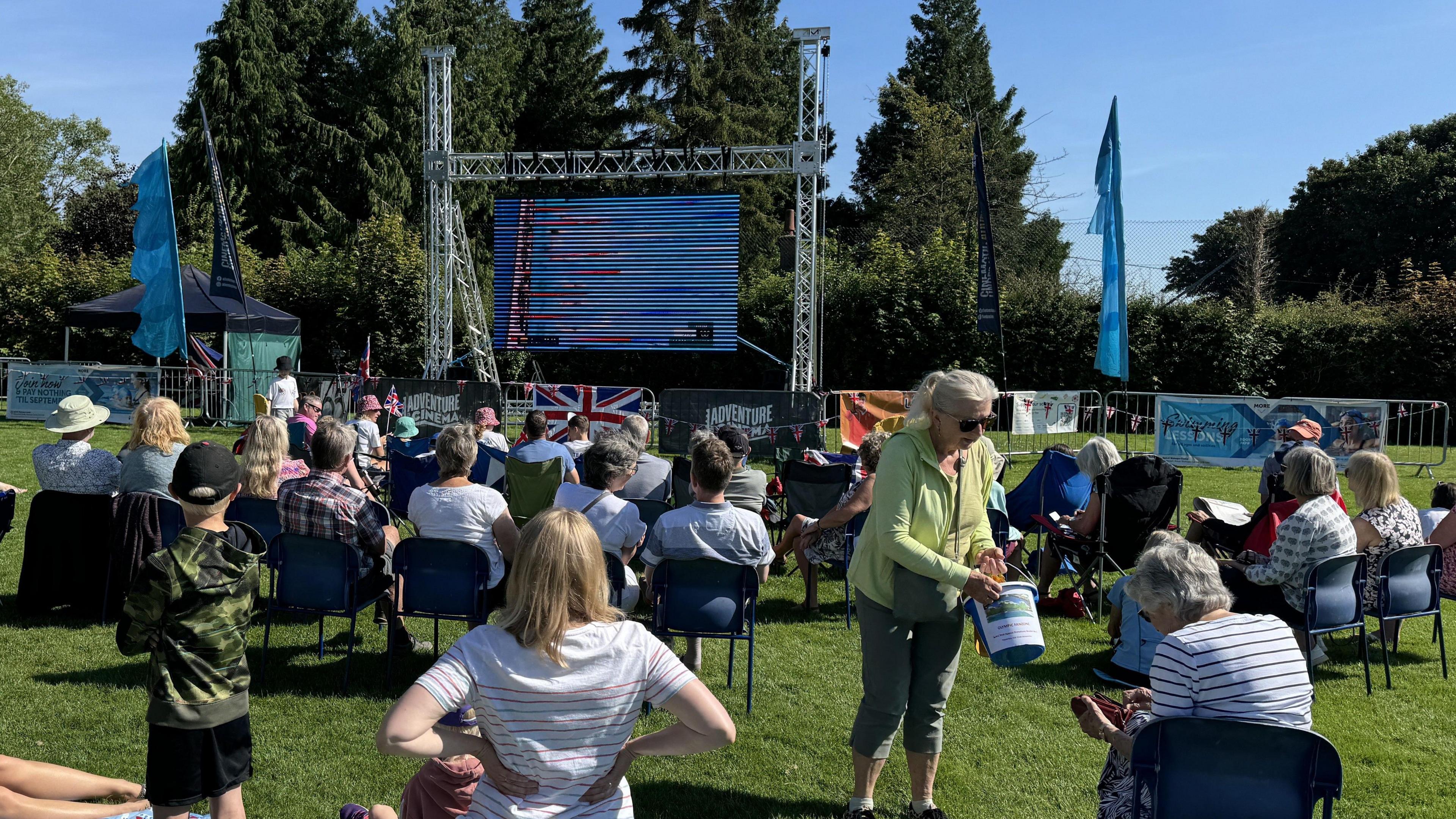 Doznes of people sit on fold-out chairs on a green field. They are watching a large screen which has been put up between metal scaffolding. A woman is holding out a fundraising bucket to people sitting down.