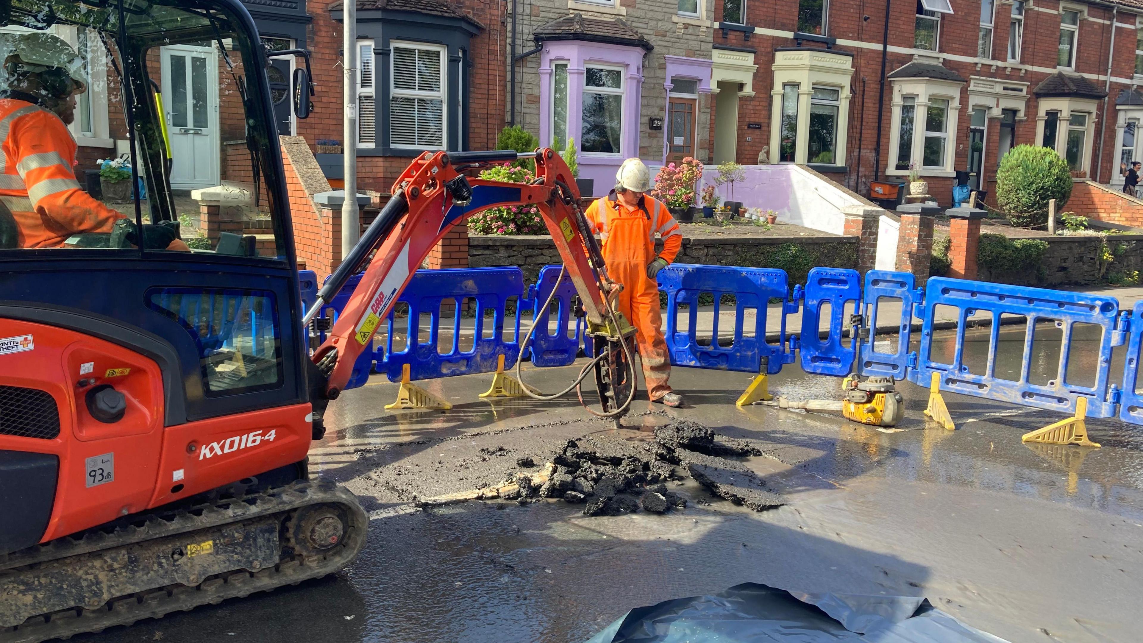 Workers are on a road where a water main has burst, which is closed in both directions. There are two people in high-visibility overalls - one inside a digger and the other standing over the hole they are working on. Behind them is a barrier to the road, and houses.