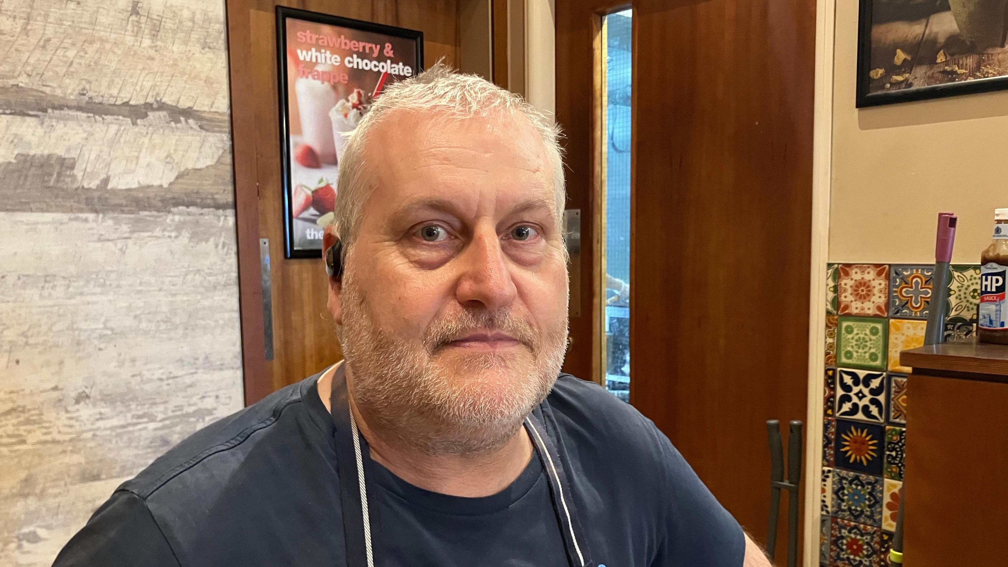 Geoff Morris is in a cafe. He has short greying hair and and beard and moustache. He is wearing a dark T-shirt and an apron