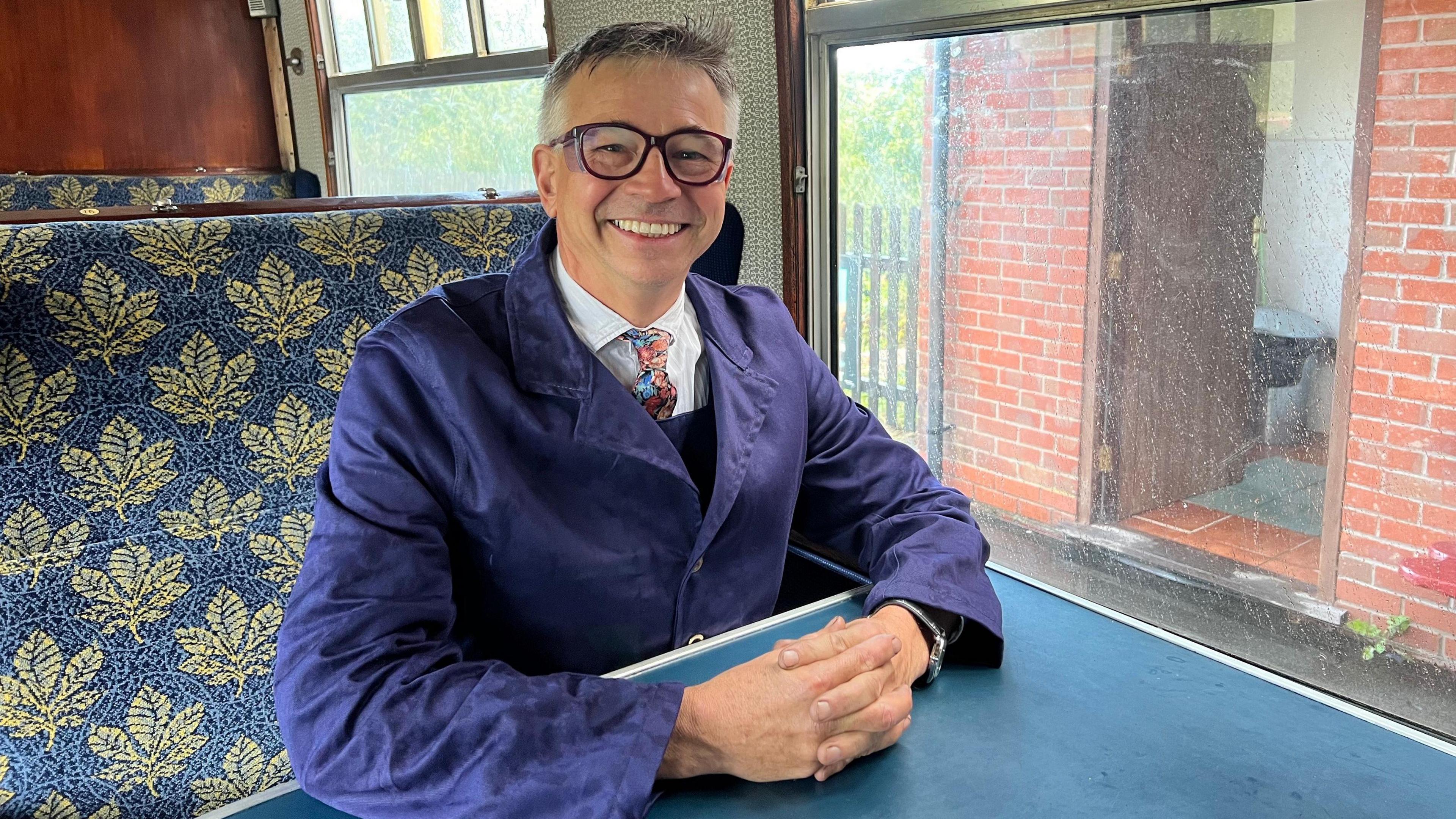 A man with glasses and grey hair, wearing a bluejacket and a tie. He's sat on a train on seats with a blue floral pattern.