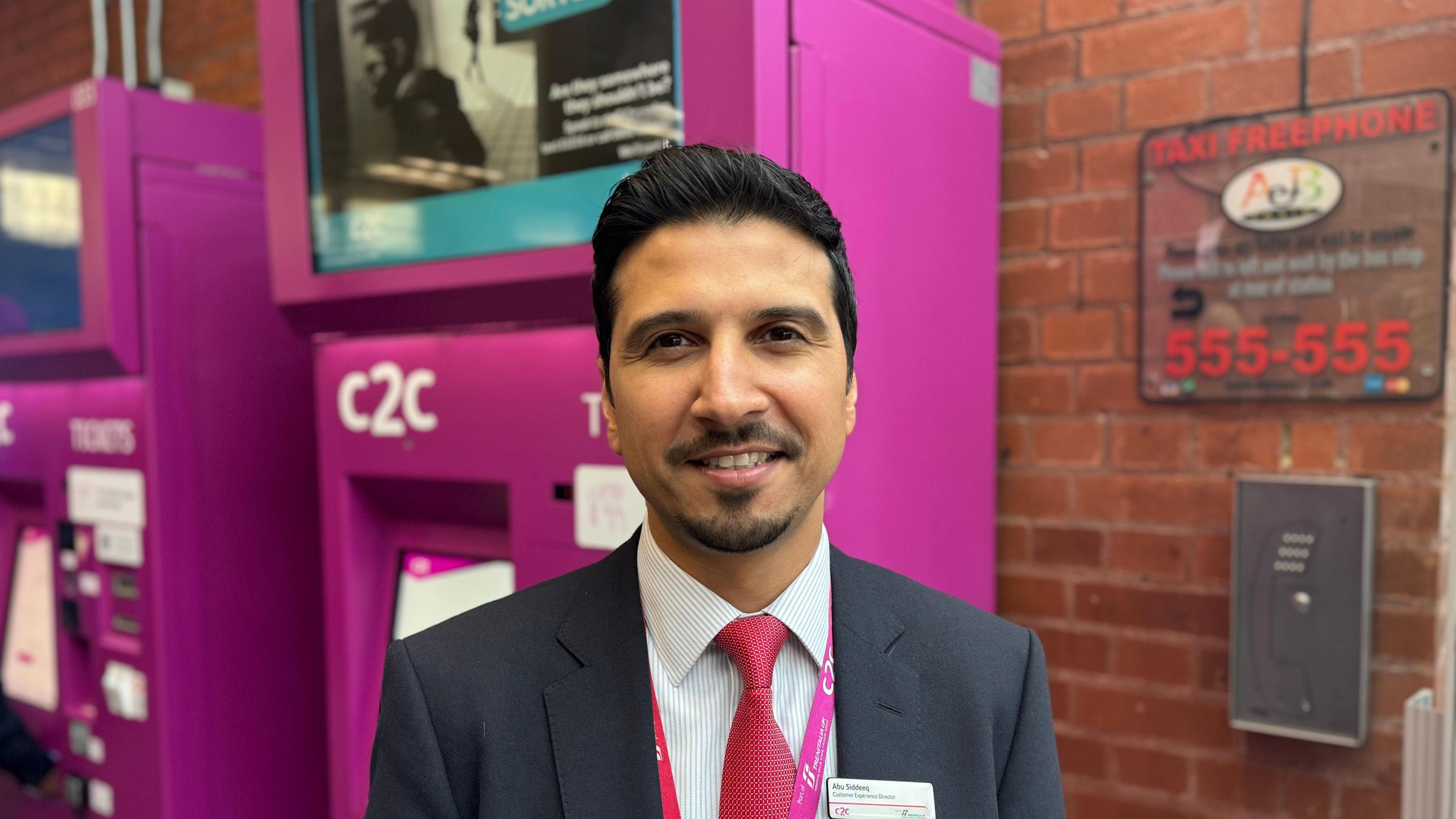 A man with short black hair and some facial hair wearing a suit standing in front of a pink ticket machine