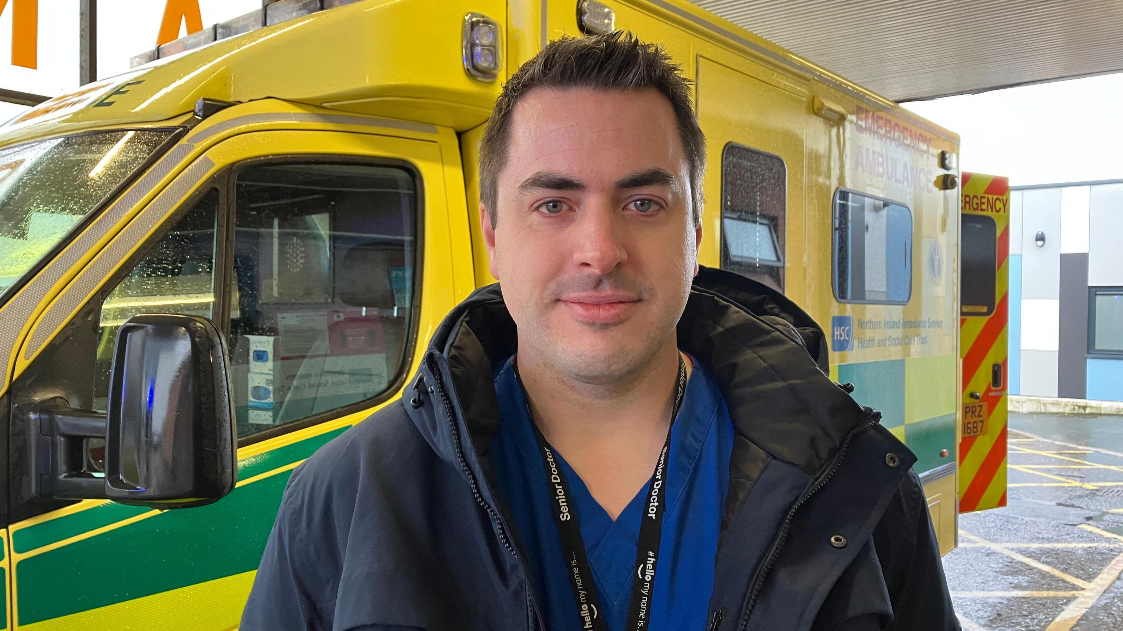 Dr Ian Dunwoody has short black hair and is standing in front of an ambulance wearing a black coat over his blue hospital scrubs. 