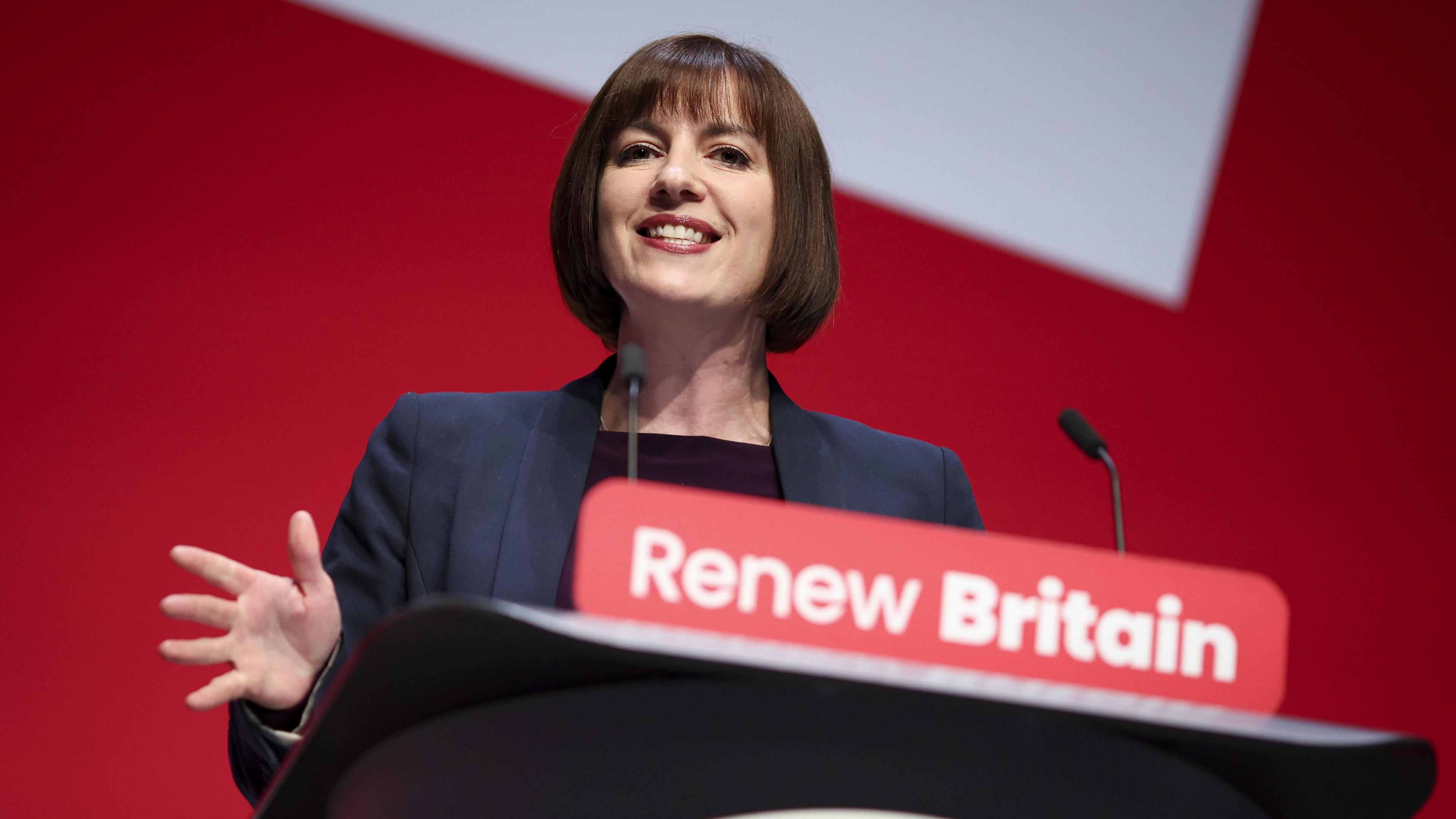 The education secretary is smiling while delivering her speech behind a podium which has "renew Britain" written on the front. She is wearing a navy blazer over a dark top.