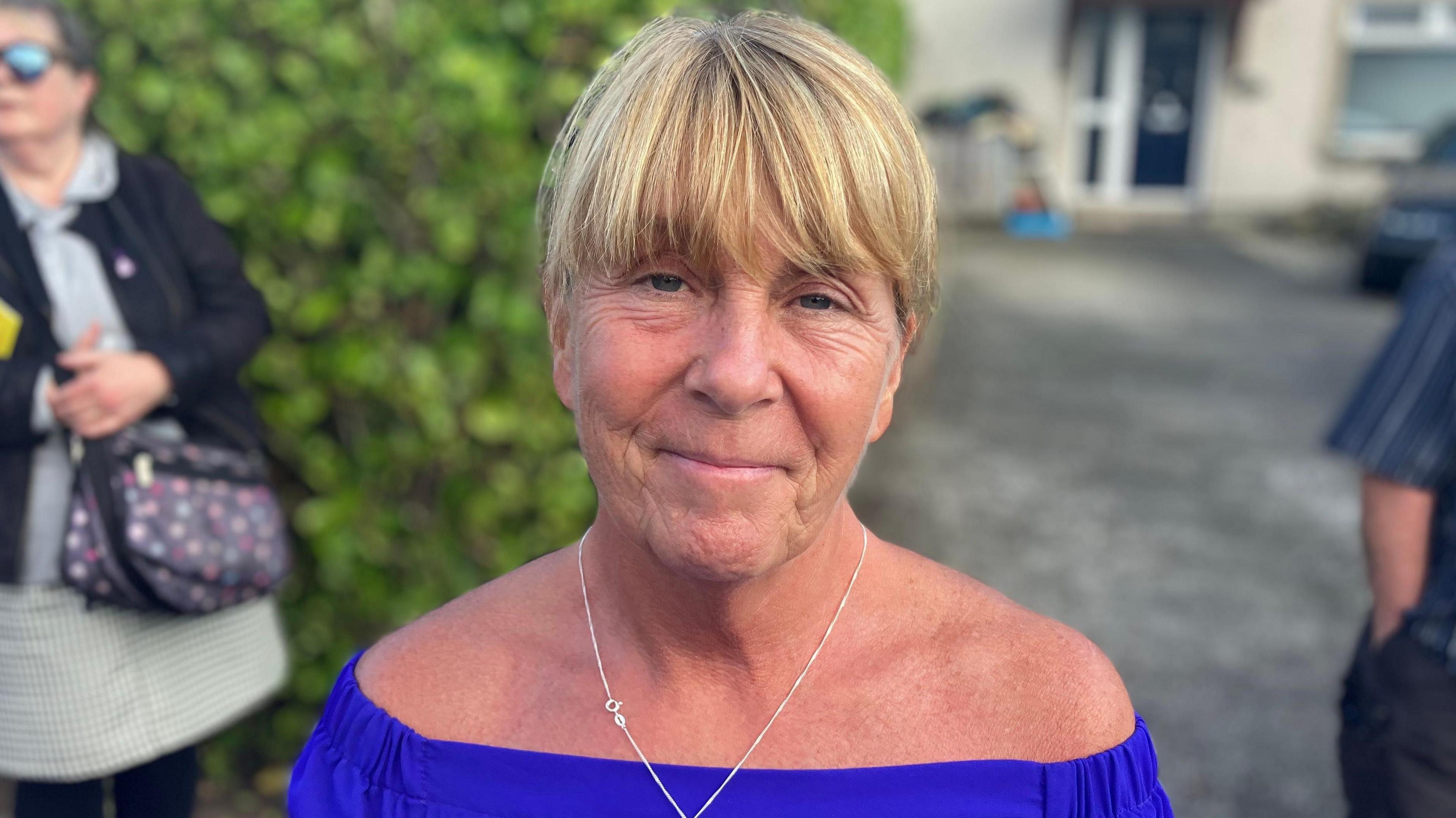 A woman is wearing a blue top with a silver chain. She has blonde hair. 