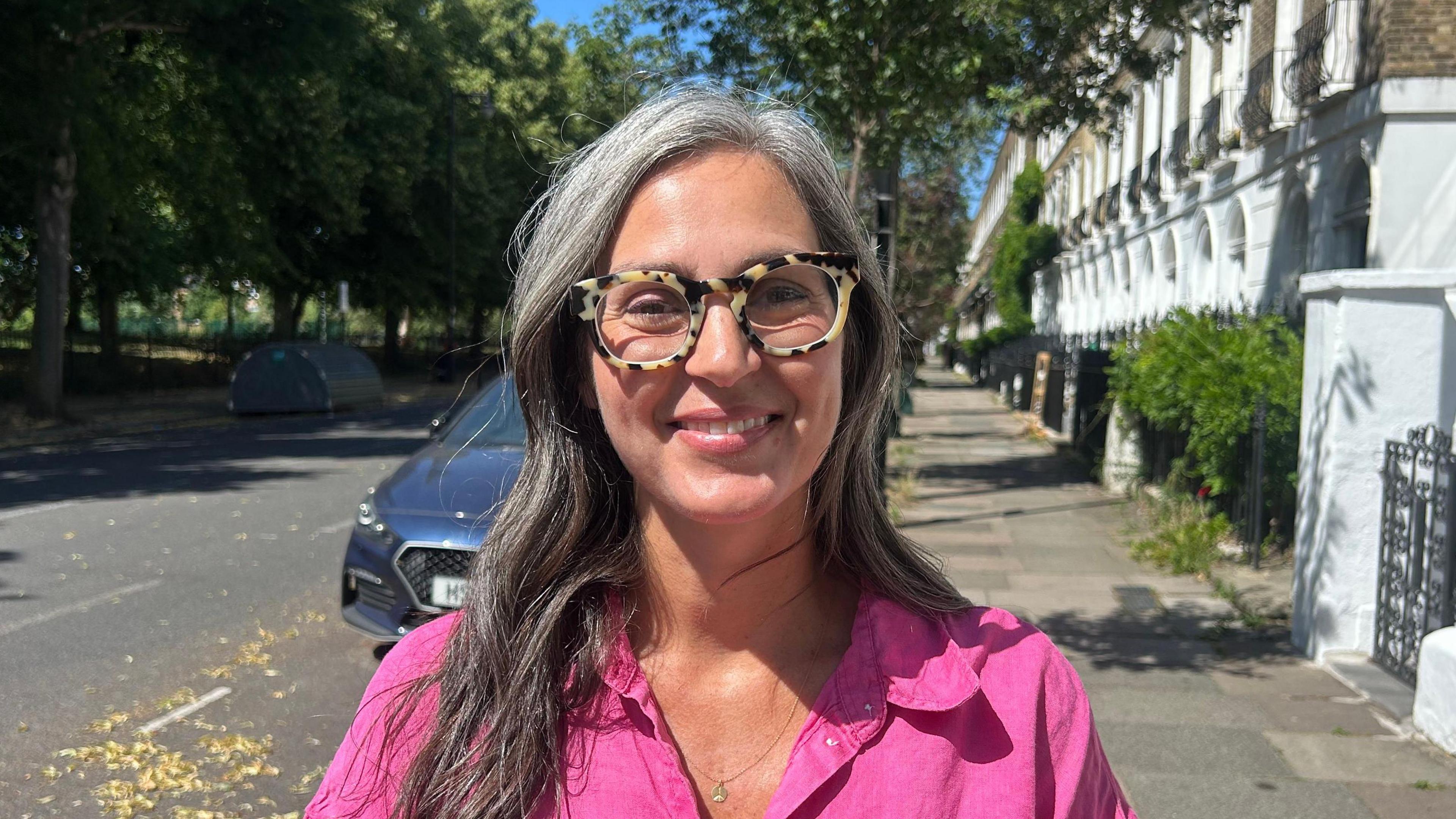 Woman with tortoiseshell framed spectacles, grey and white long straight hair, and a bright pink shirt stood on a pavement in a residential area. She smiles at the camera.