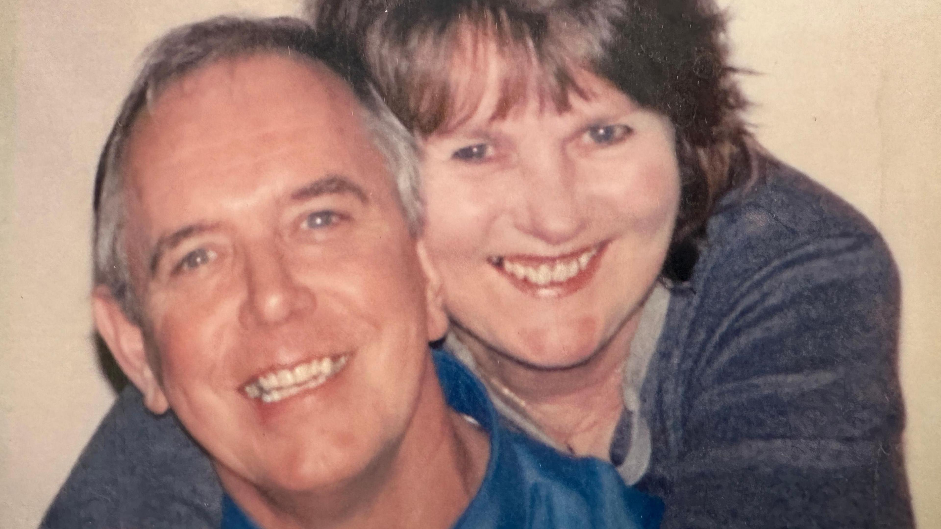 This is a close up photo of Mel and June smiling to camera. Mel is in the foreground and June is behind him with her arms around him. 