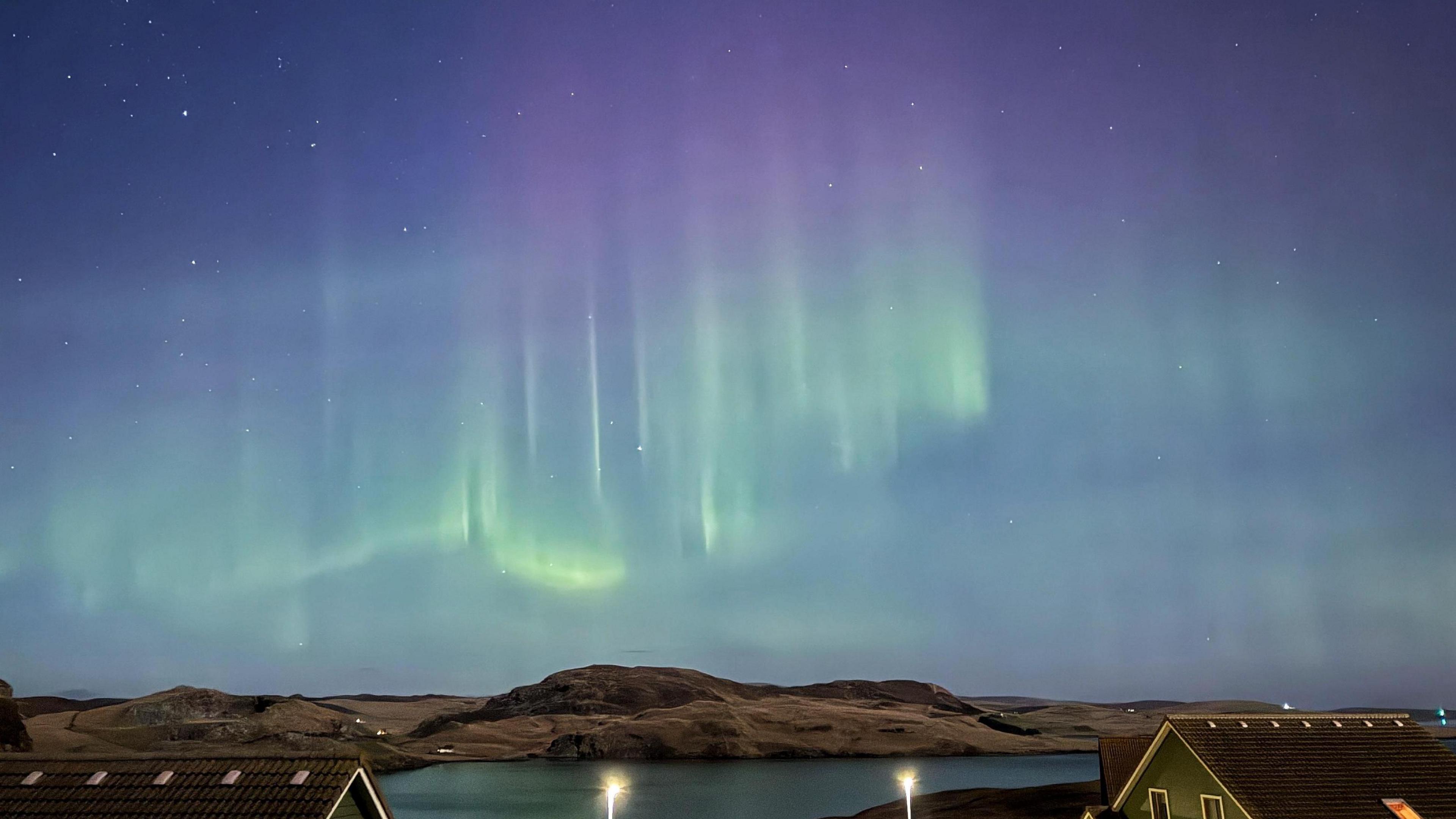 Bright green aurora and a hint of purple light shimmer in a blue evening sky over water and hills with houses and street lights in the foreground