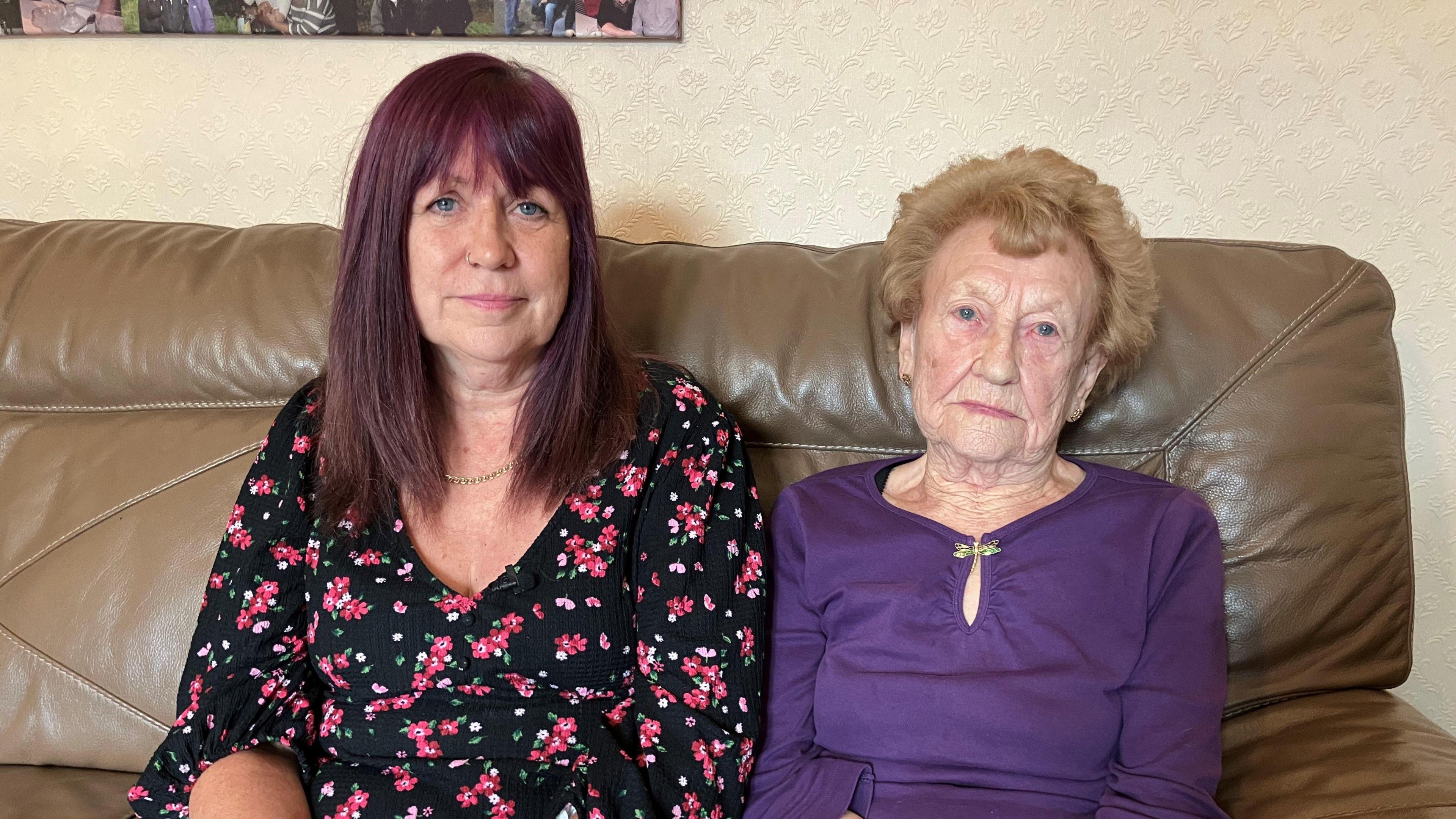 A woman with dark pink hair wearing a pink and black floral top, sat to the left of an older woman wearing a purple top. They are both sat on a brown leather sofa and are looking forward.
