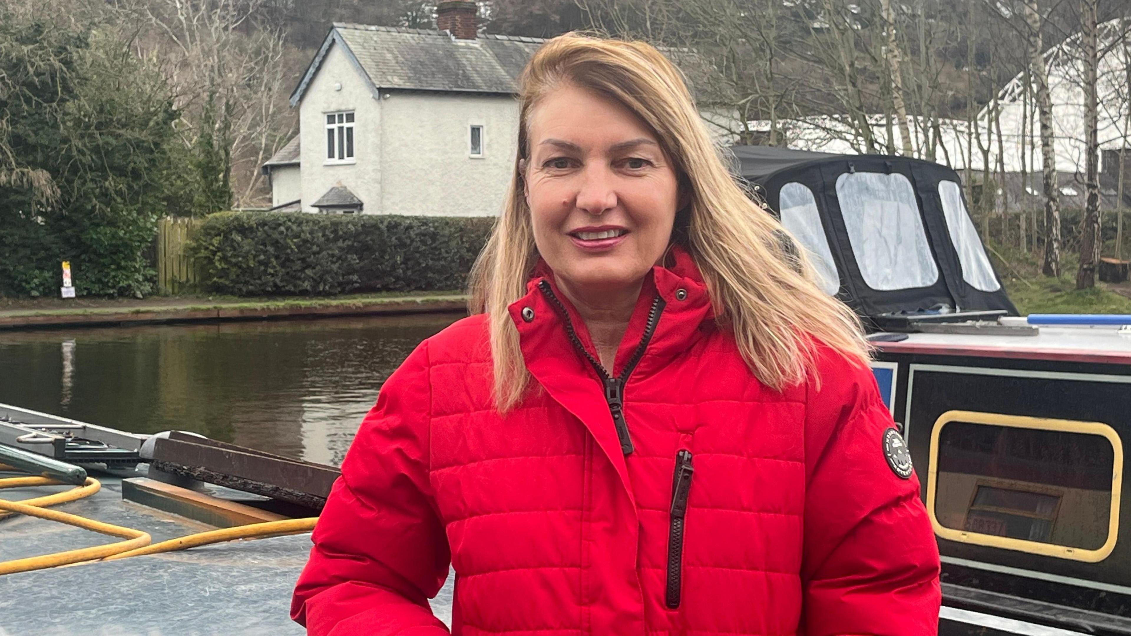 Llun menyw gwallt melyn sy'n gwisgo cot goch yn sefyll o flaen cwch ar gamlas