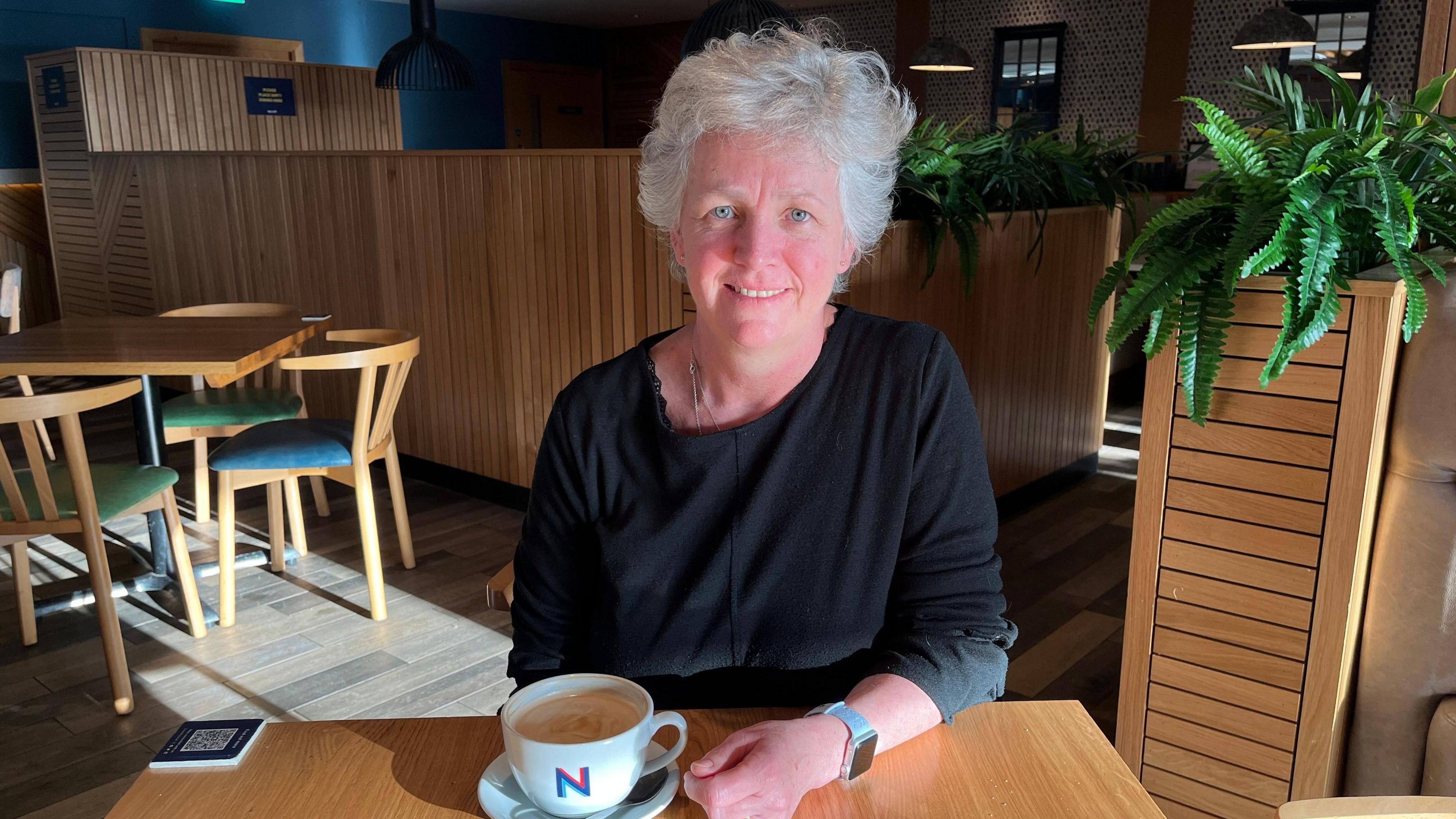 A woman with short grey hair and wearing a black top and grey watch is sitting at a table with a cup of coffee in a white cup.  There is another table and chairs behind the woman.