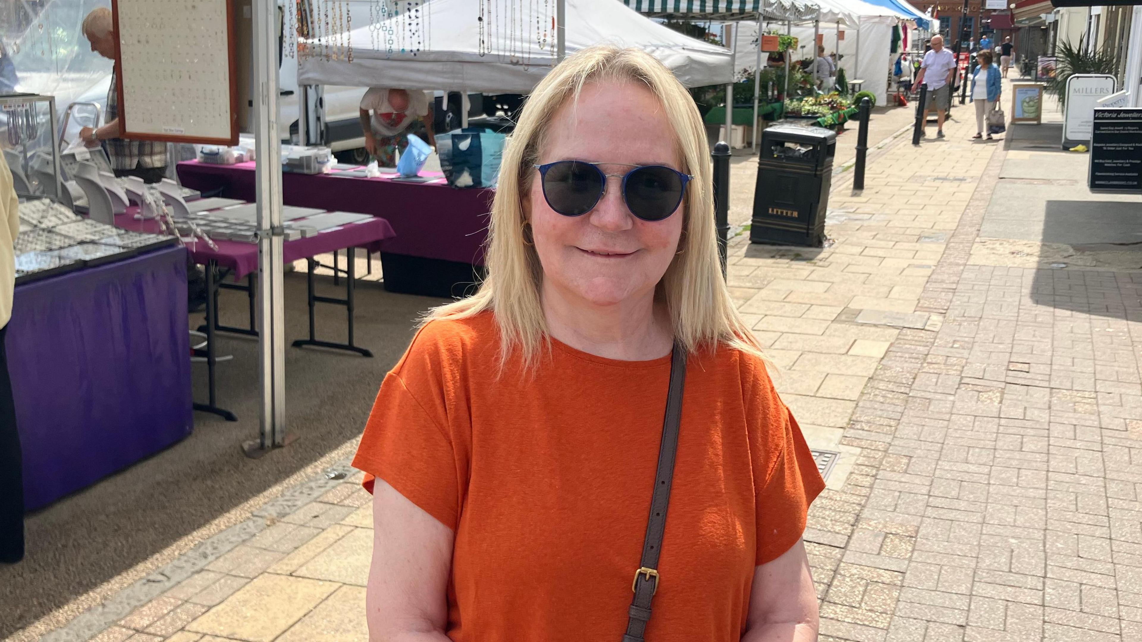 Barbara Frost is looking at the camera and is wearing an orange T-shirt. She has should-length blonde hair and is wearing dark glasses. There are market stalls and people shopping in the background