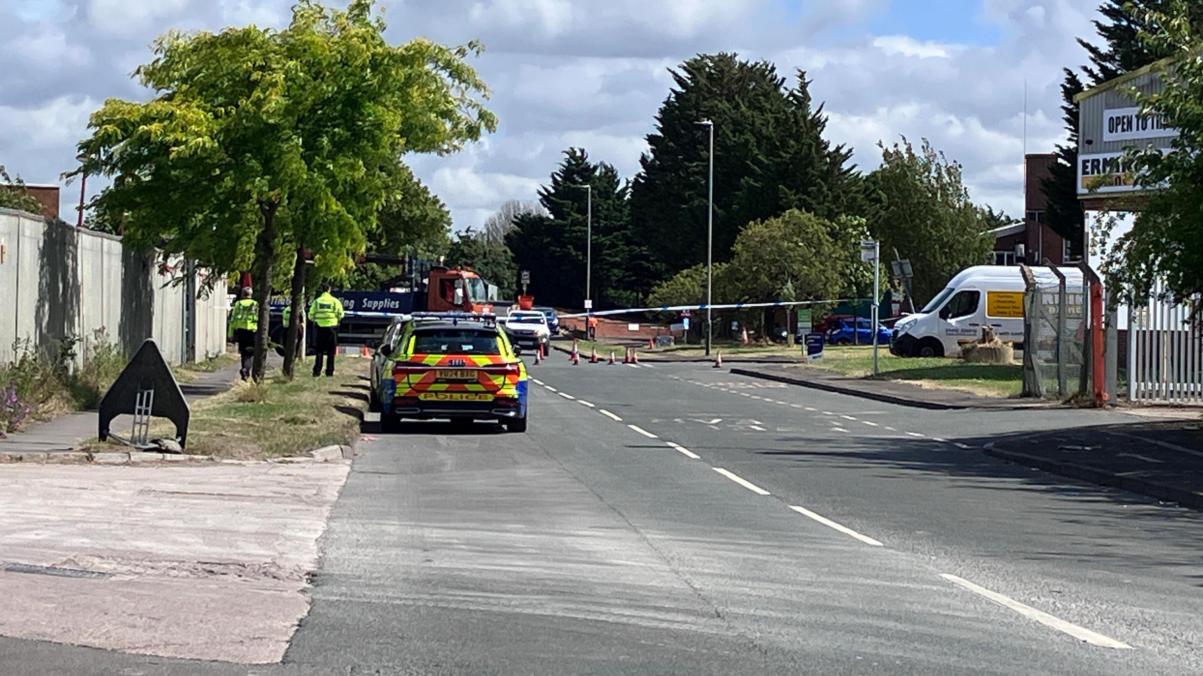 The rear view of a police car with an orange and yellow fluorescent boot door, parked on a road closed with white and blue police tape with police officers walking on the pavement in the distance where a lorry can be seen parked across the road