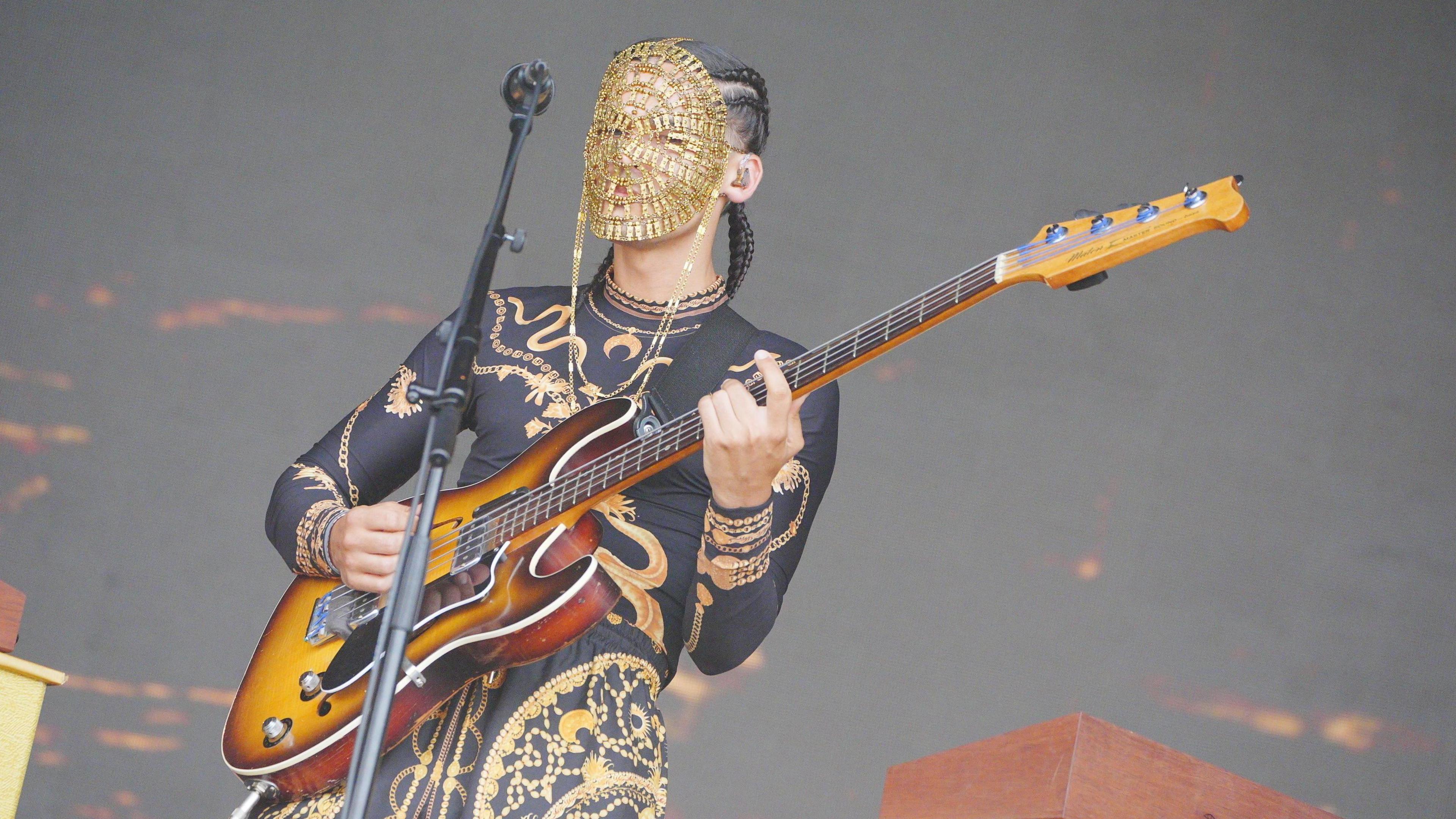 A performer in a black and golden dress playing the electric guitar in front of a microphone. They are wearing a golden mask, obscuring their face.