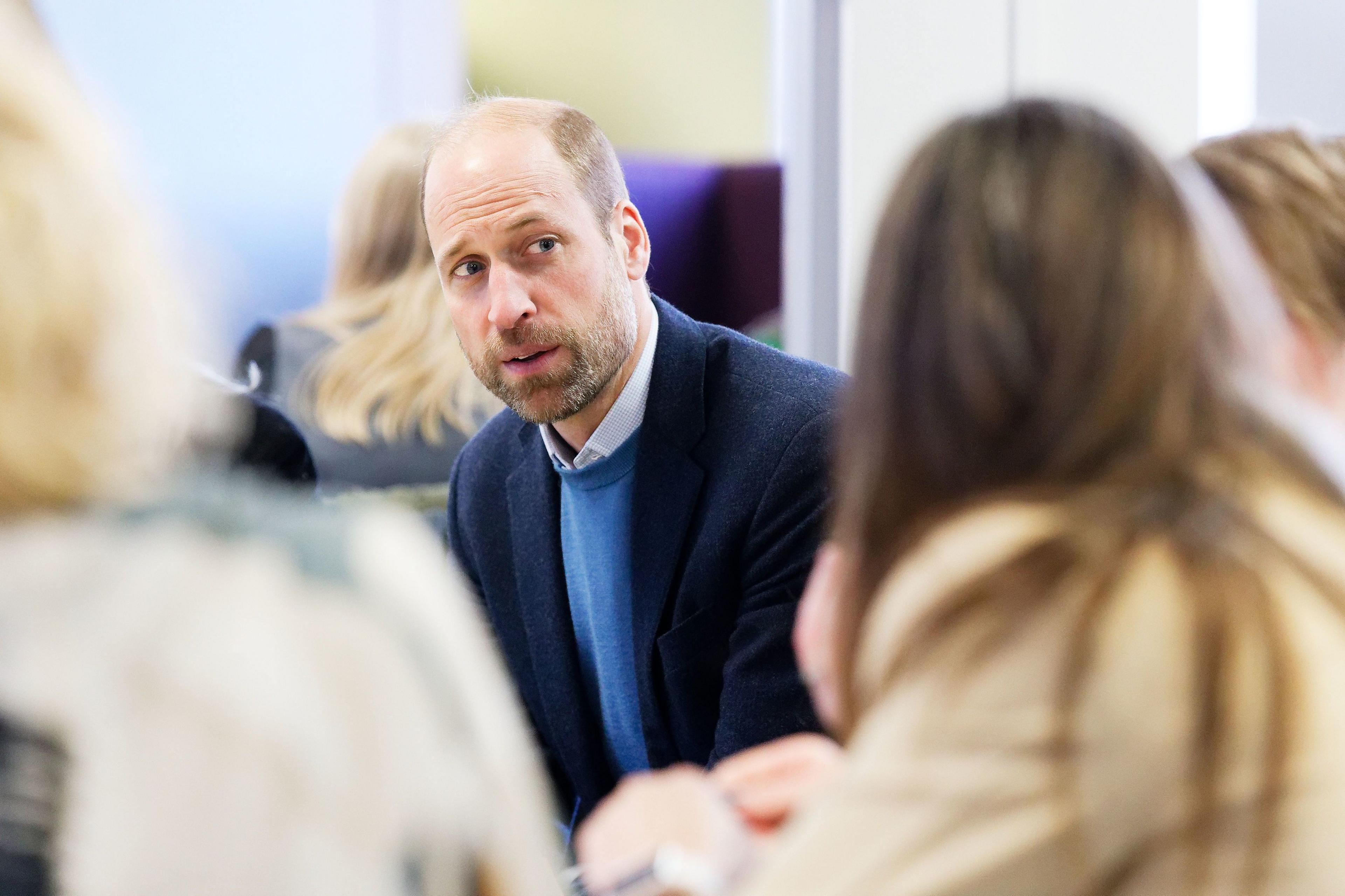 Prince Wililam, wearing a light blue jumper and darker jacket, is seen speaking to a group of women 