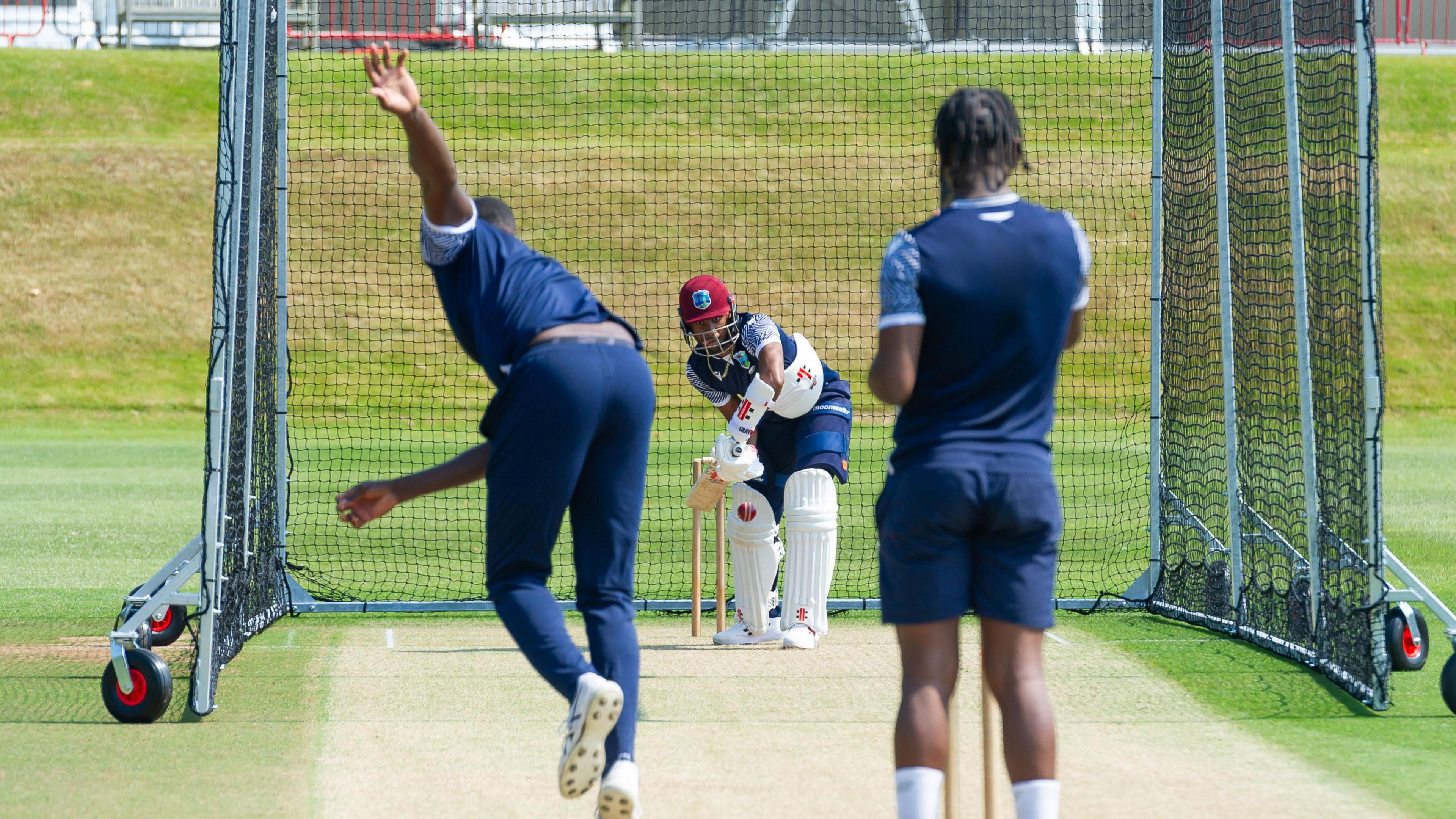 Three people in a cricket net. Two people are wearing blue and facing away. One is holding a bat waiting to hit the ball.