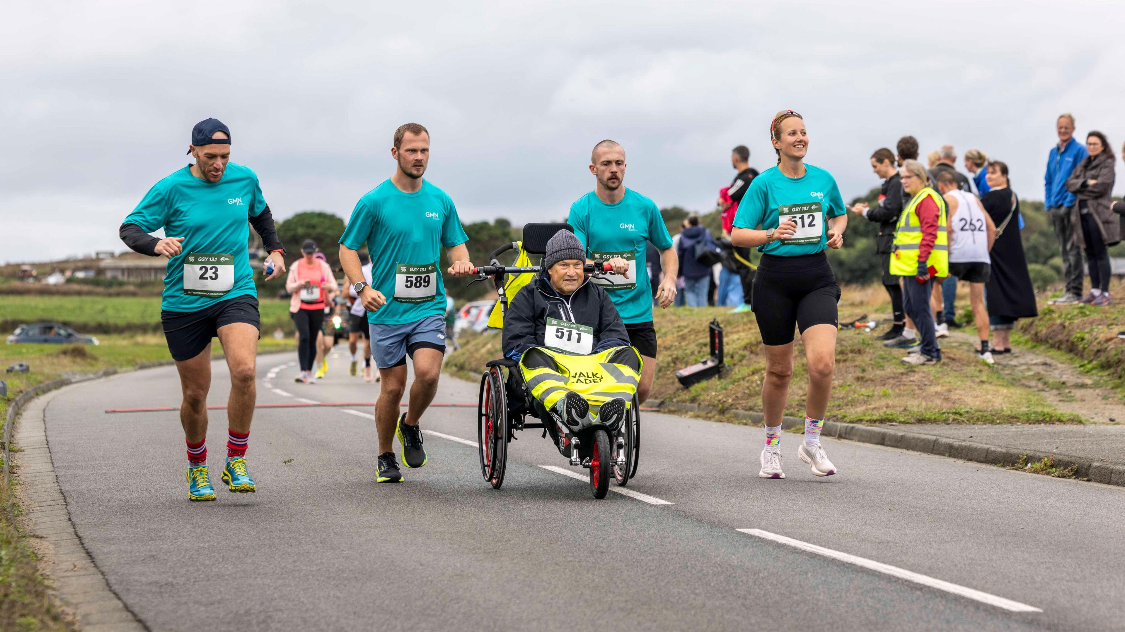 Four runners in matching teal tops run along a country road, watched by spectators. Two of them push a man in a sports wheelchair.