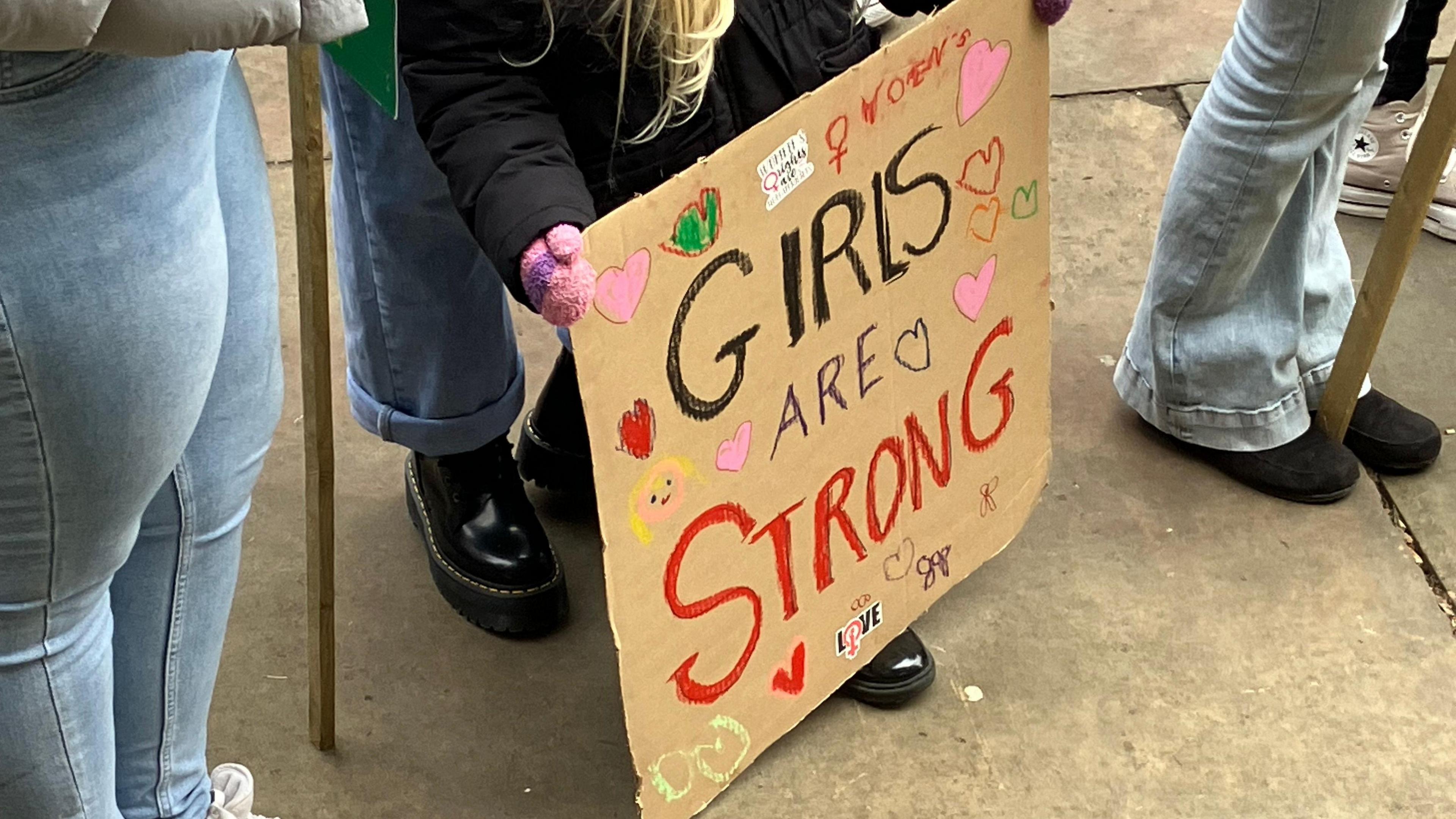 A girl holds a cardboard sign with the words 'girls are strong' written across it. Her face isn't visible.