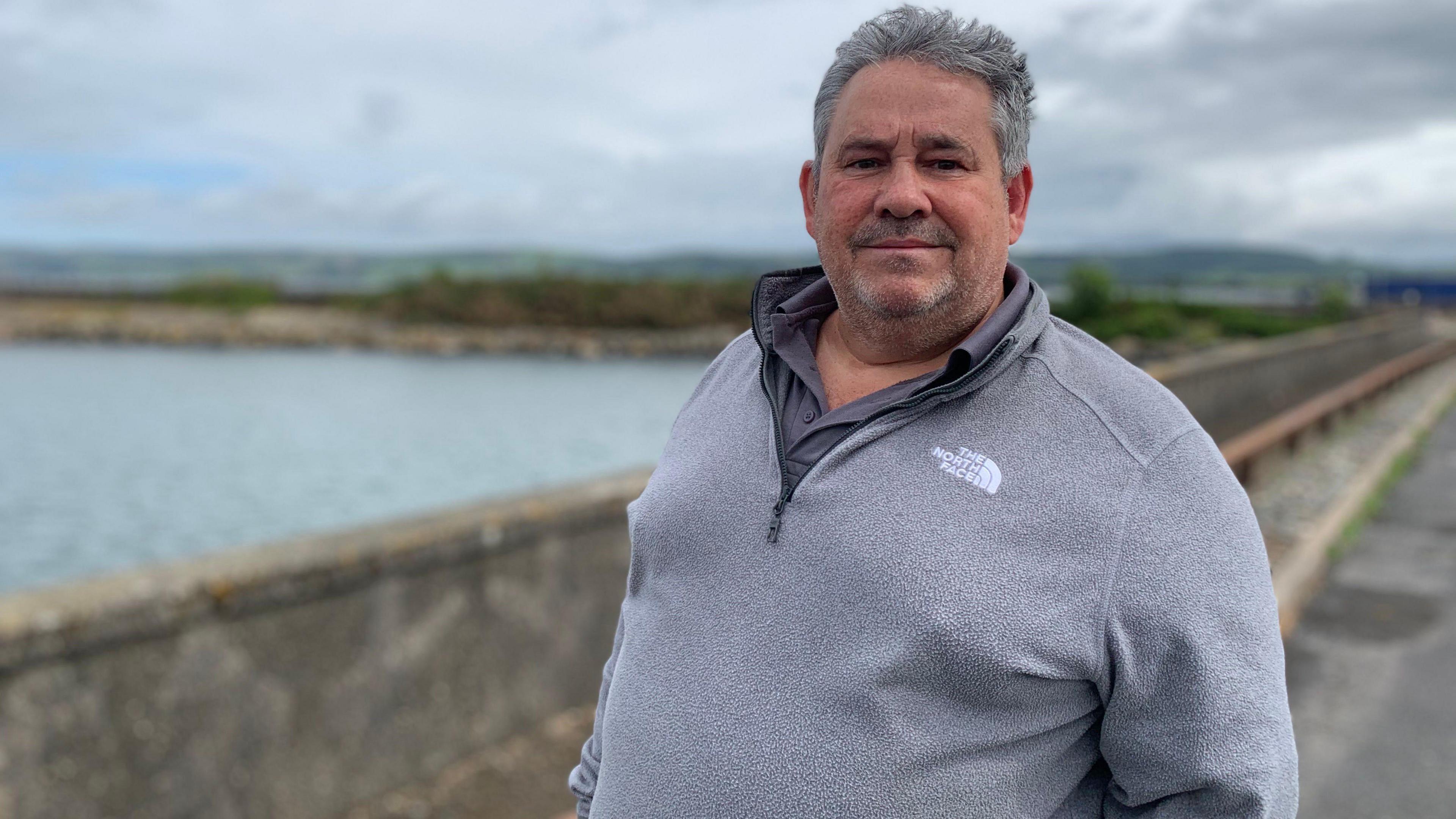 A man with grey hair in a grey fleecy top stands in front of the harbour wall in Stranraer