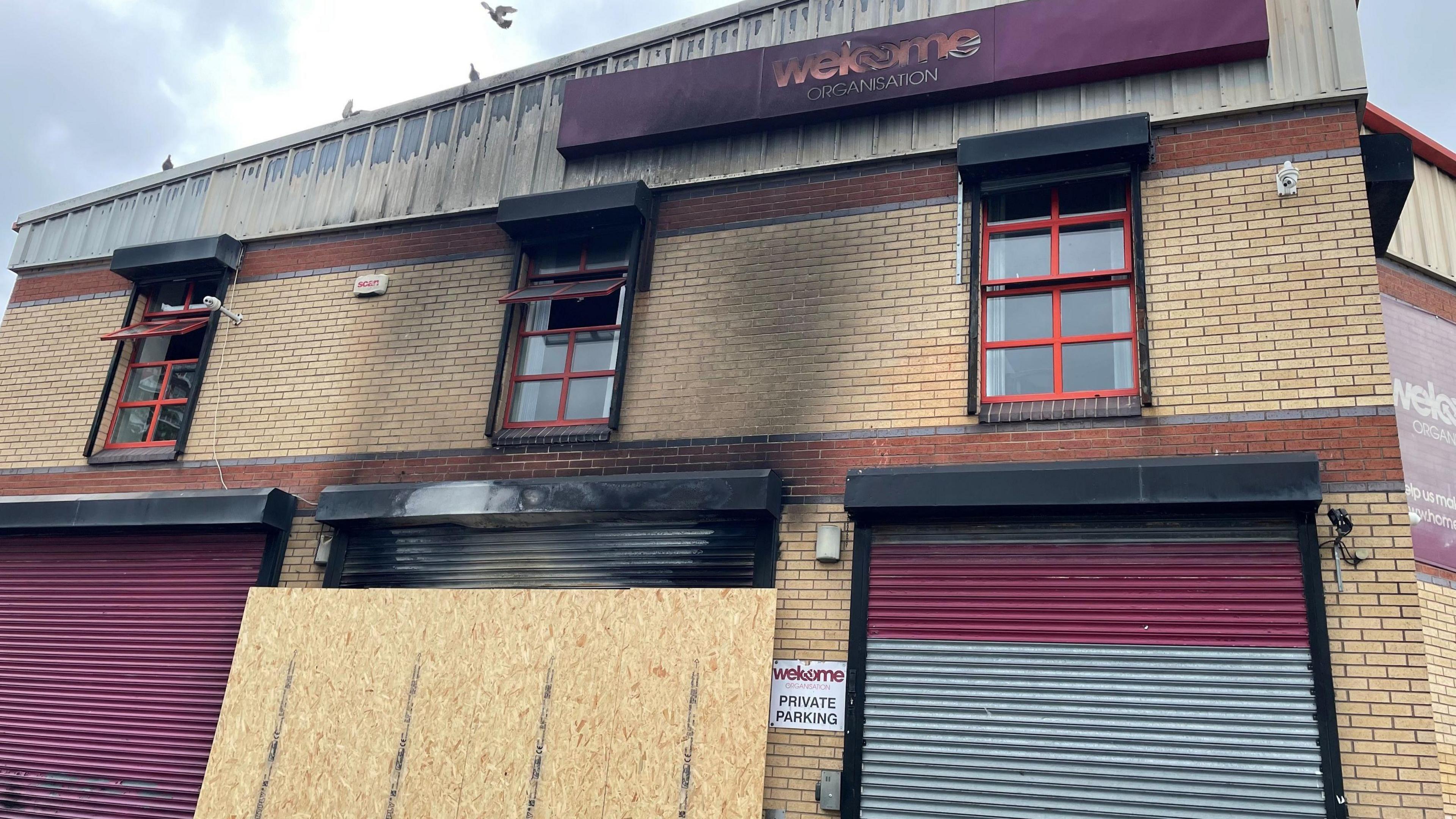 The damaged Welcome Organisation HQ with a boarded-up shutter and smoke damage