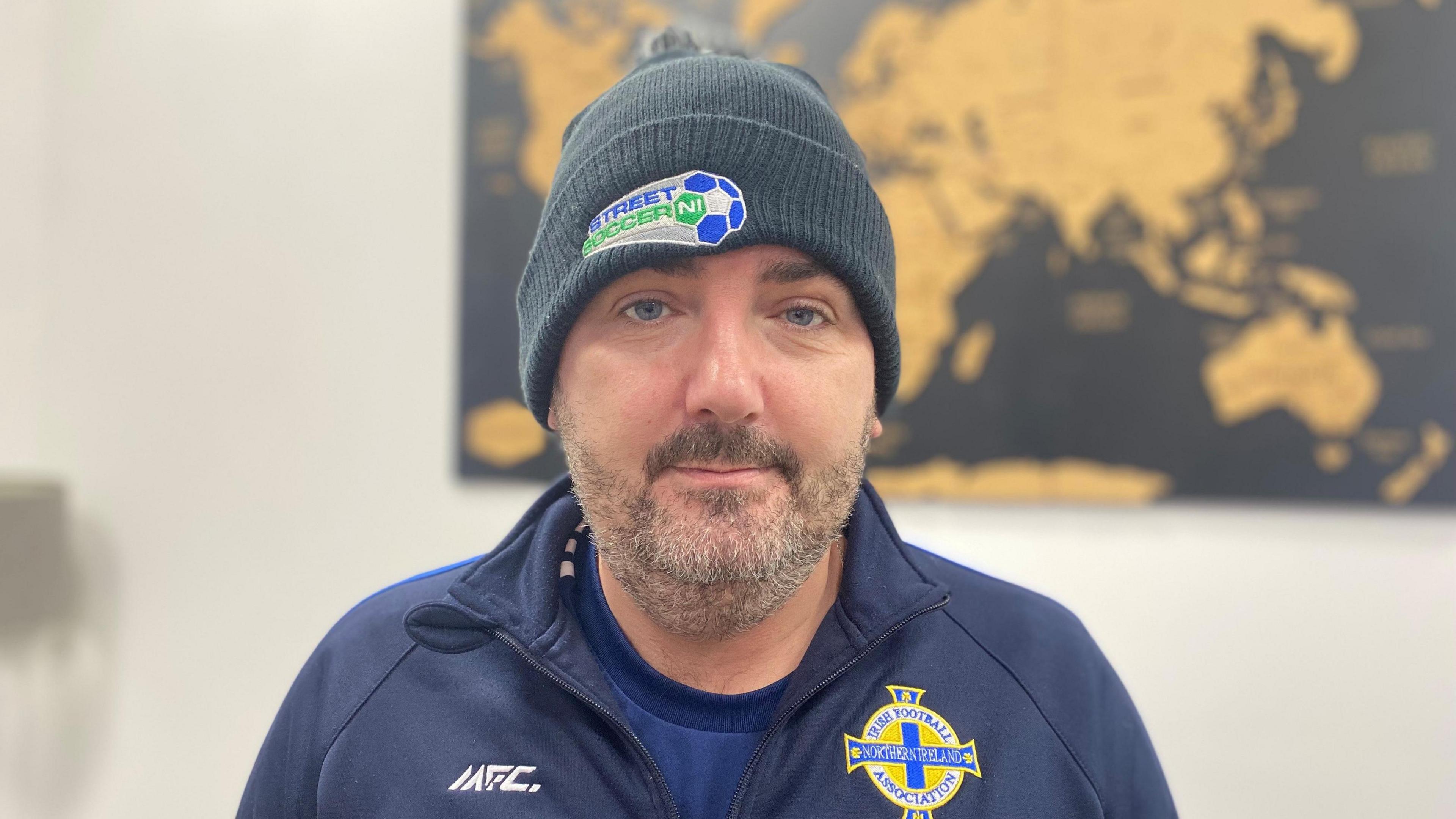 Wilbur is looking at the camera and is wearing a Street Soccer NI branded hat. He has stubble and wearing a Northern Ireland football training jacket. 