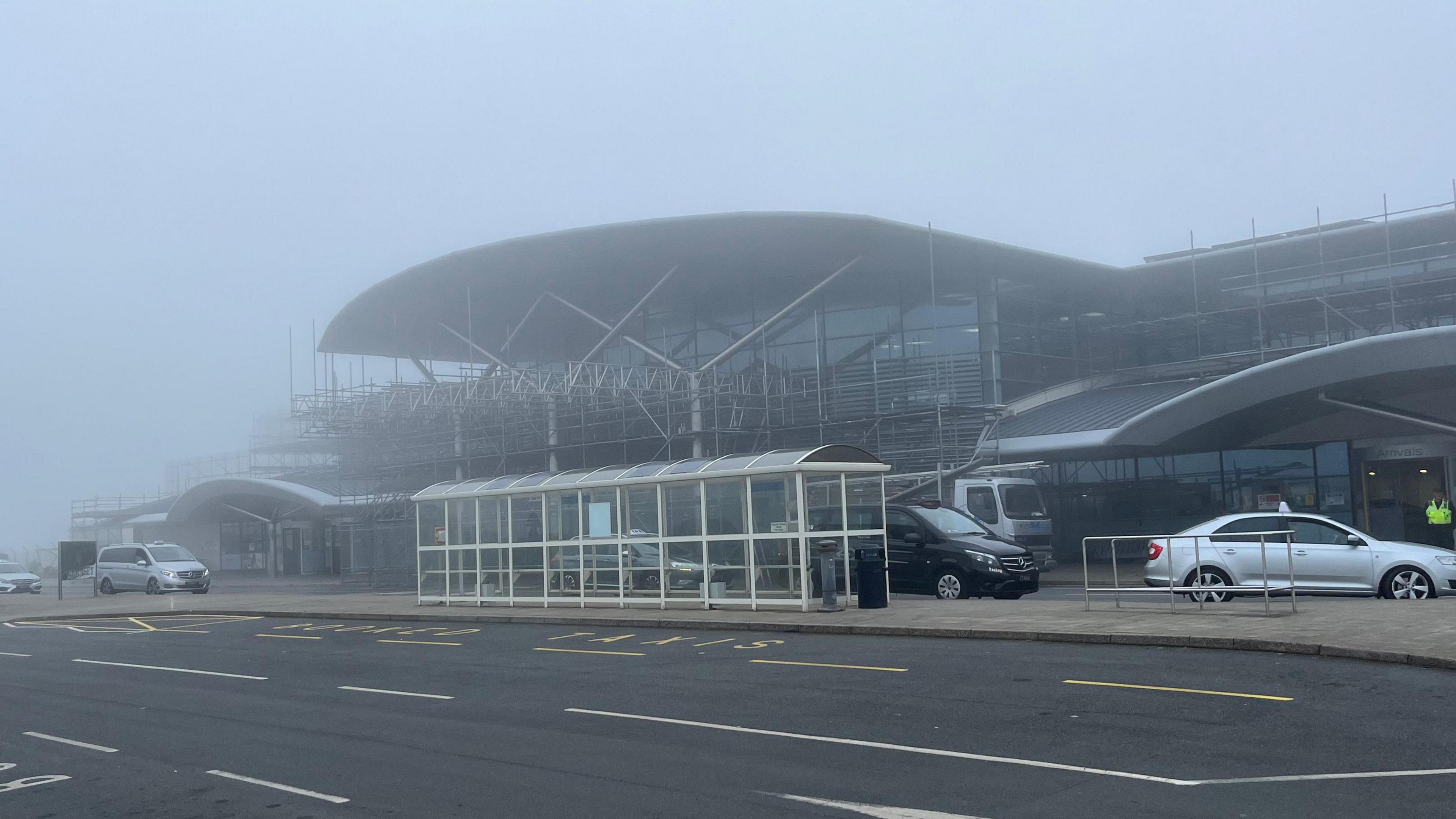 Airport buildings in glass and steel and a bus stop with a dark van and a truck and a car in fog and a man in a hi vis jacket standing in the entrance to the airport.