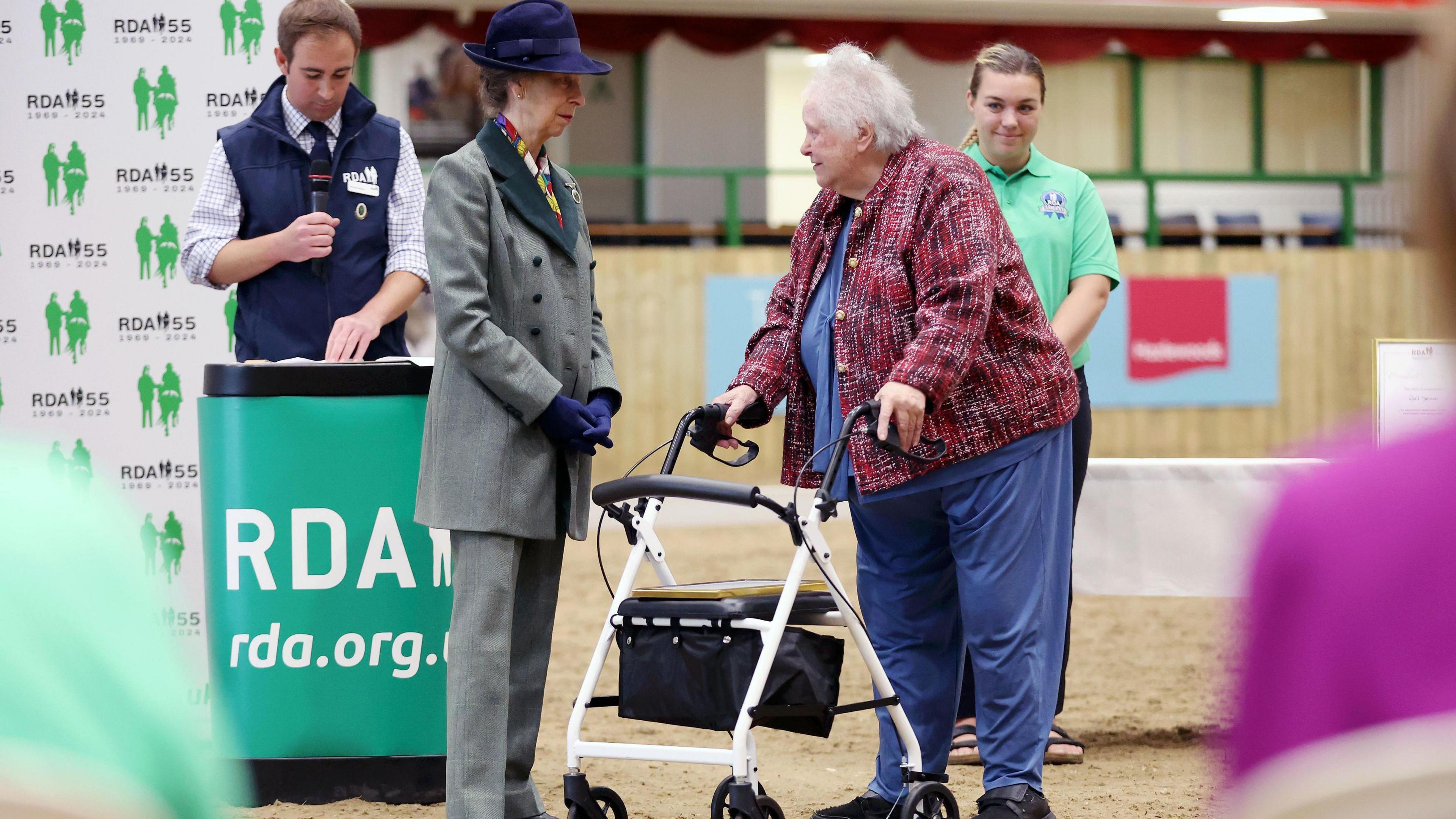 Princess Anne meeting a woman. Princess Anne is wearing a green jacket and trousers, and gloves and a hat. The woman is wearing blue trousers and a top, with a red jacket. 