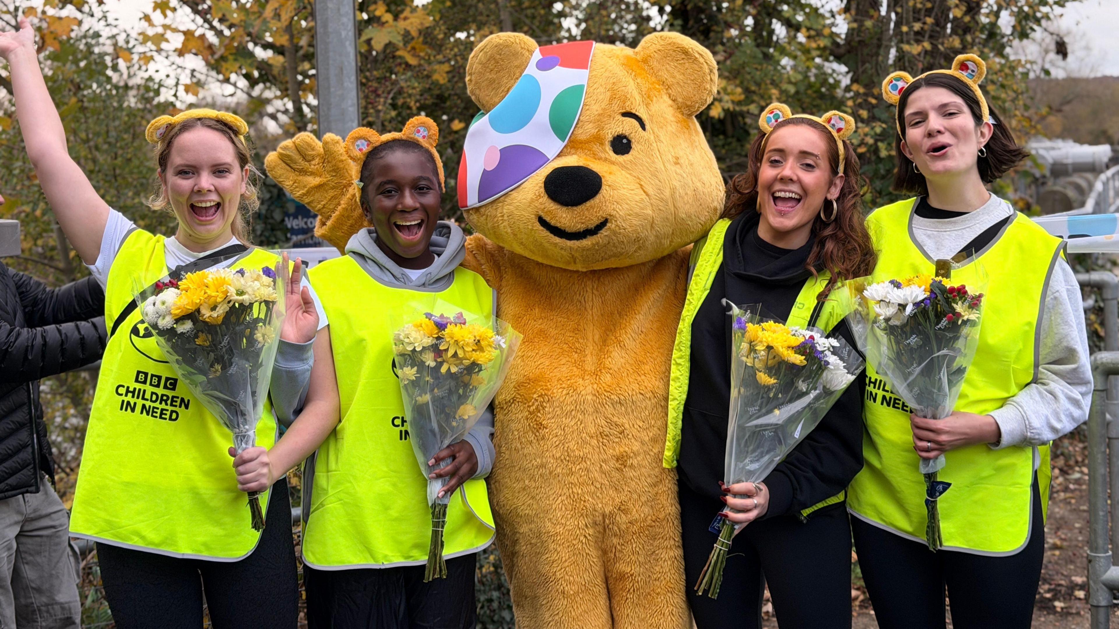 Sophie Drew, Kemi Spence, Pudsey, Ellie Tutt and Katie Tyler. The presenters are wearing high visibility vests and holding bunches of flowers while cheering.