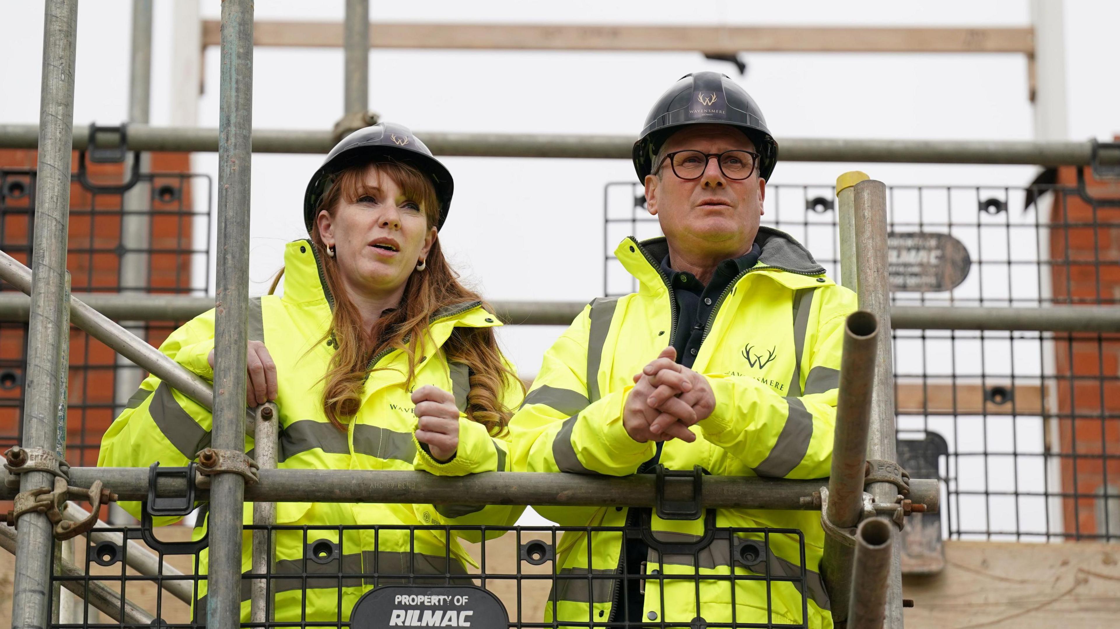 Sir Keir Starmer and Angela Rayner wearing hard hats and hi-visability jackets as they lean on scaffolding, looking out across a construction site for a housing development