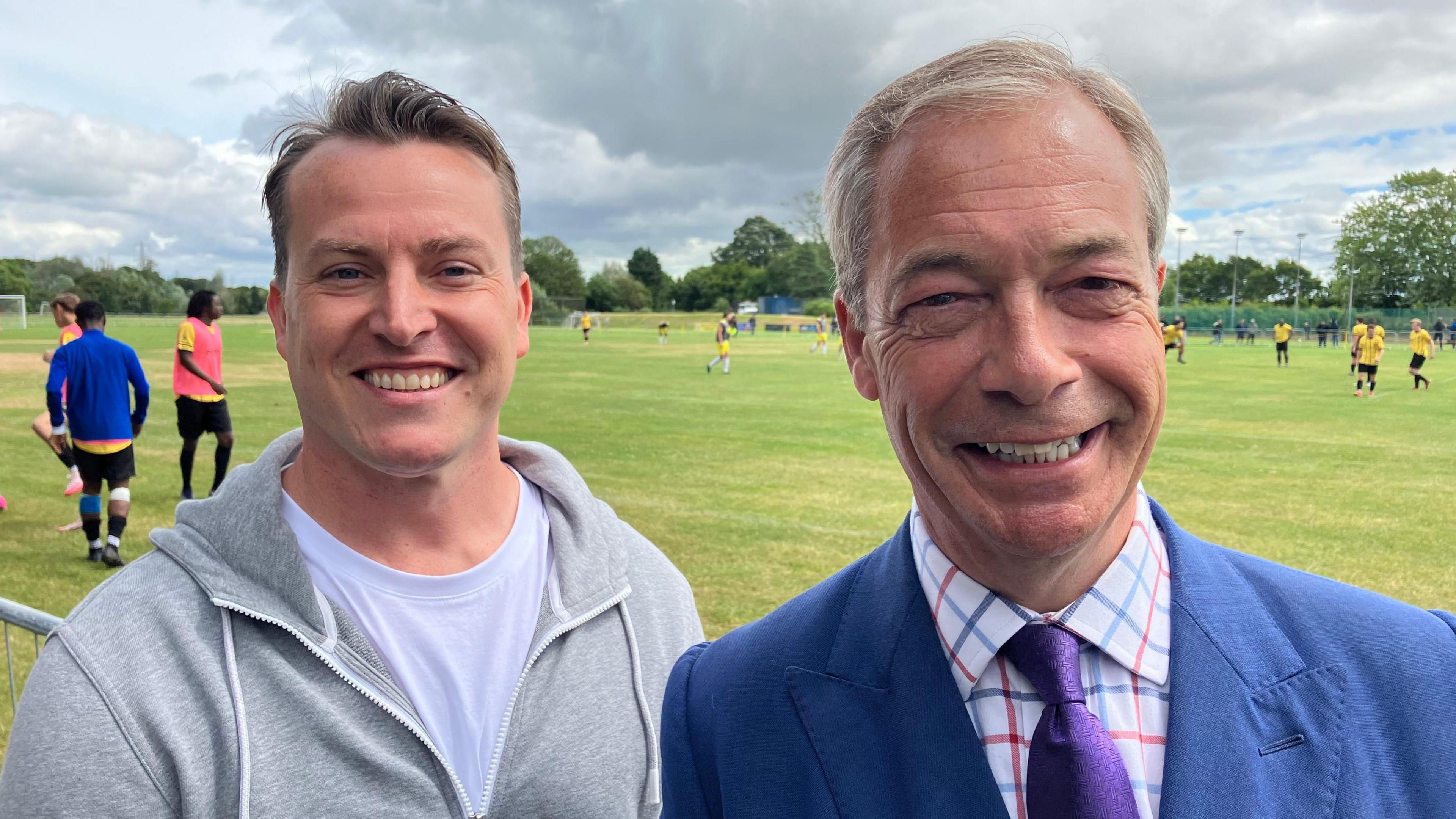 James McMurdock and Nigel Farage smile and look directly into the camera as they stand by the side of a football match. 