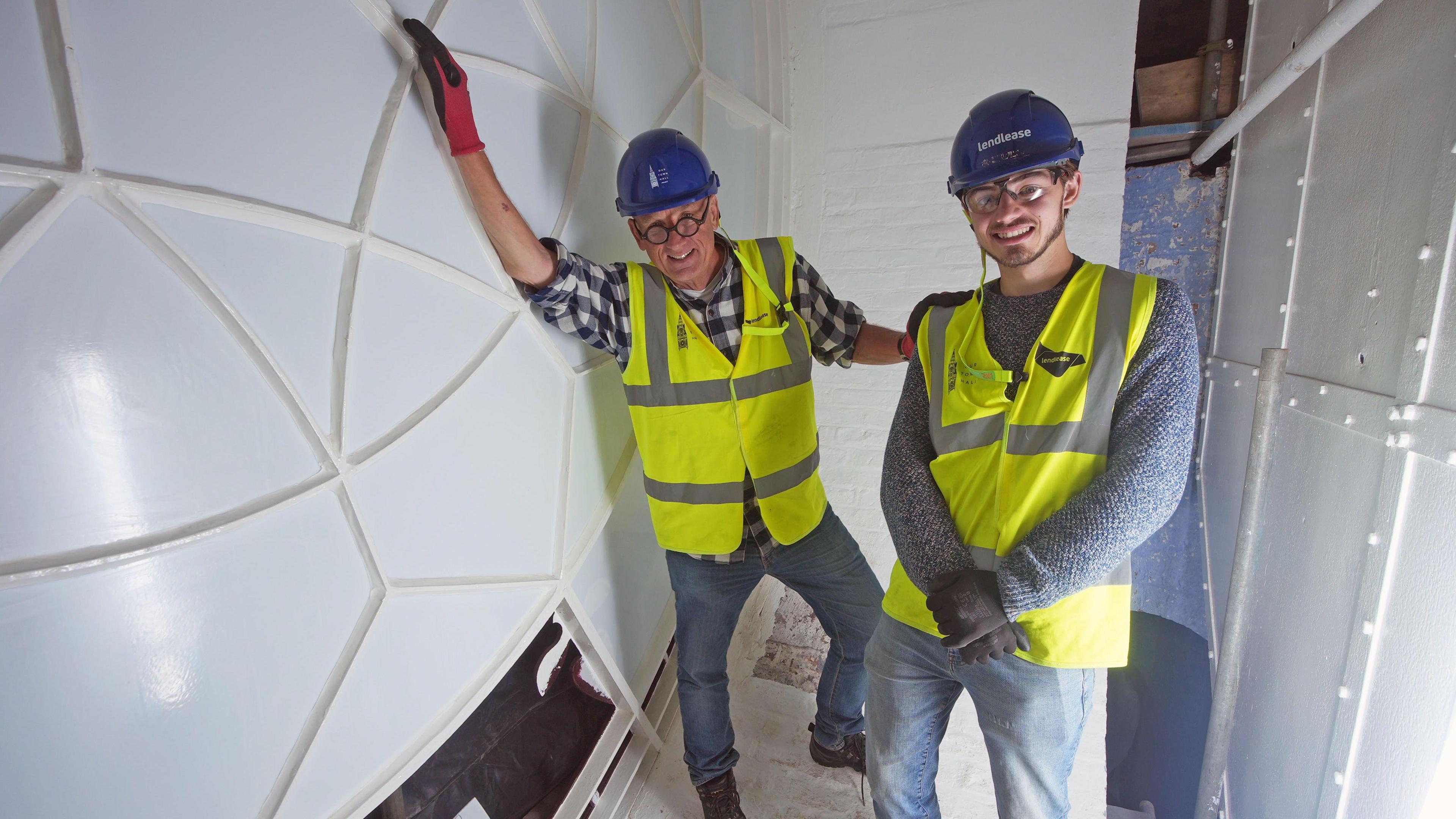 Repair shop host Steve Fletcher and horologist Mark Crangle stand on the inside of the town hall's clock face. Steve leans on a white structure with edges patterned along the outside. 