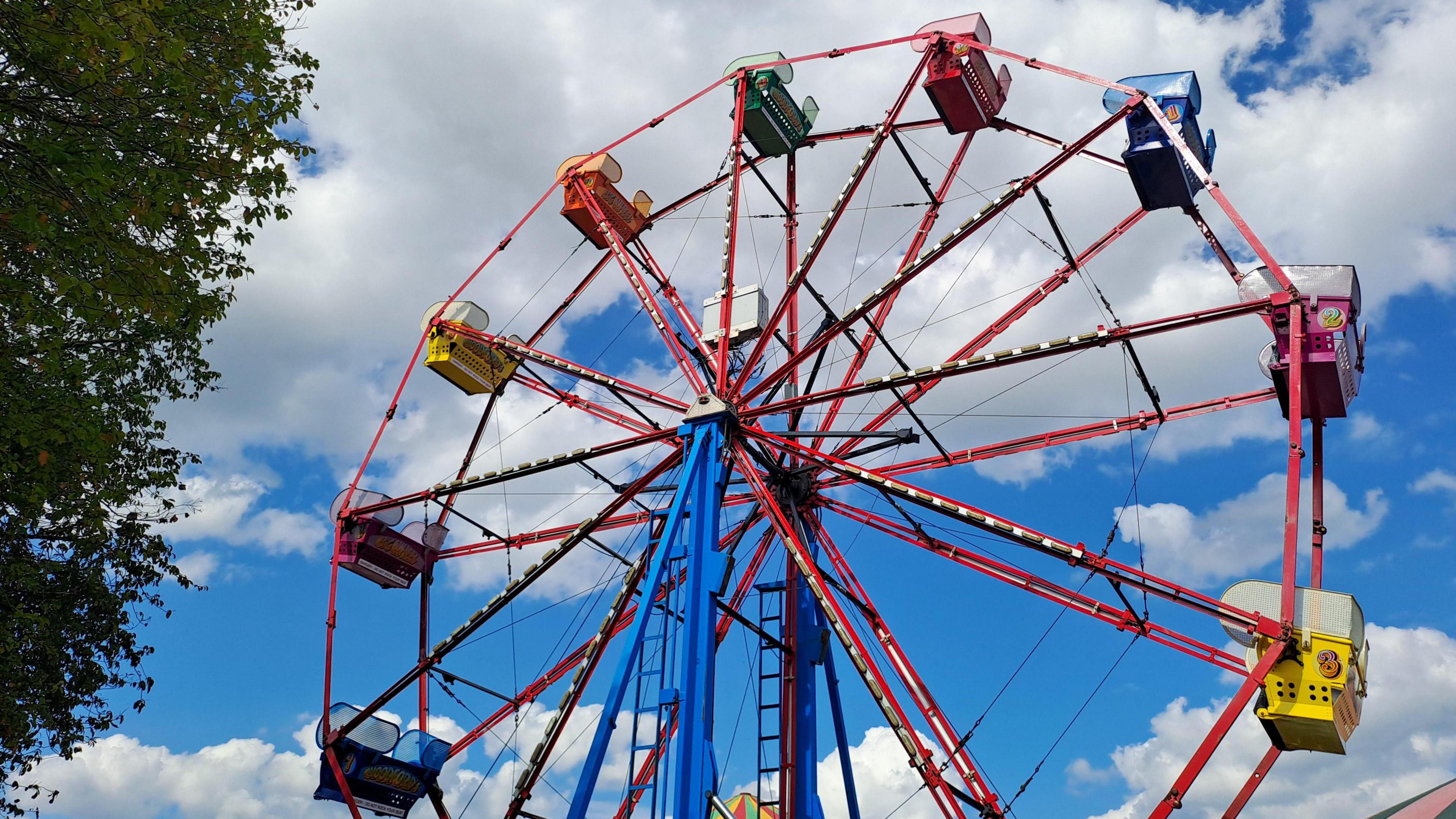 A blue sky and fluffy clouds provide the backdrop for this image of a bright coloured ferris wheel with different coloured seats