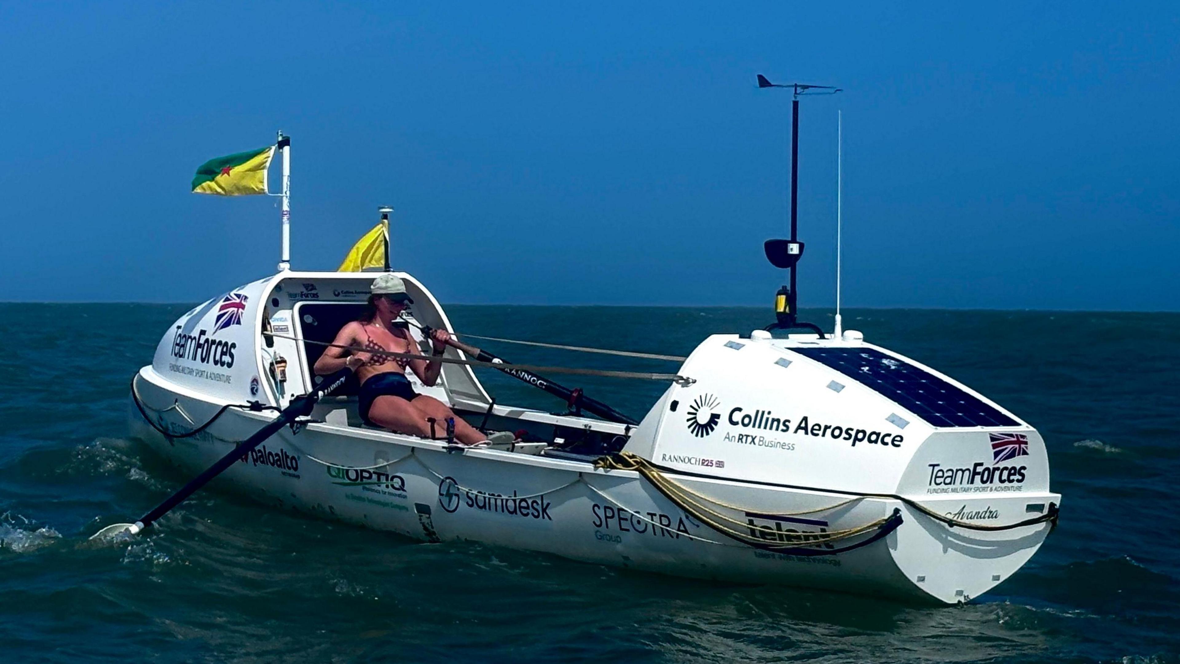 Zara is rowing her boat on the sea. The sky is empty and light blue, the sea is also clear but a much darker blue. The is a line where the two appear to meet. Zara is rowing her boat and wears a baseball cap, bikini top and shorts. She looks slightly sunburnt and there is no shelter above her.  