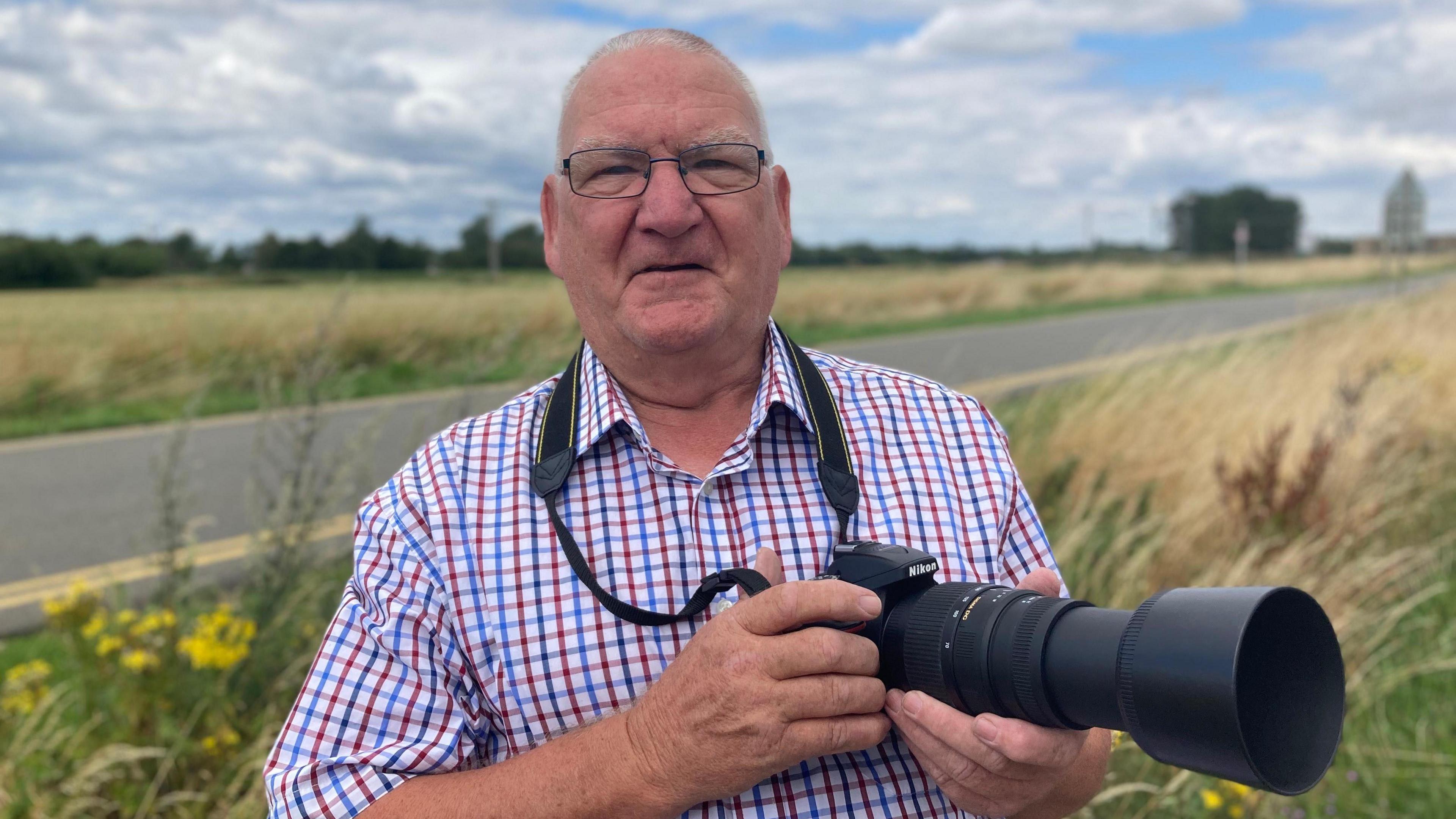 David Fyfe, wearing glasses and checked shirt, holding a black long-lens camera. He is smiling as he stands on an overgrown grass verge beside a road under a cloudy blue sky.