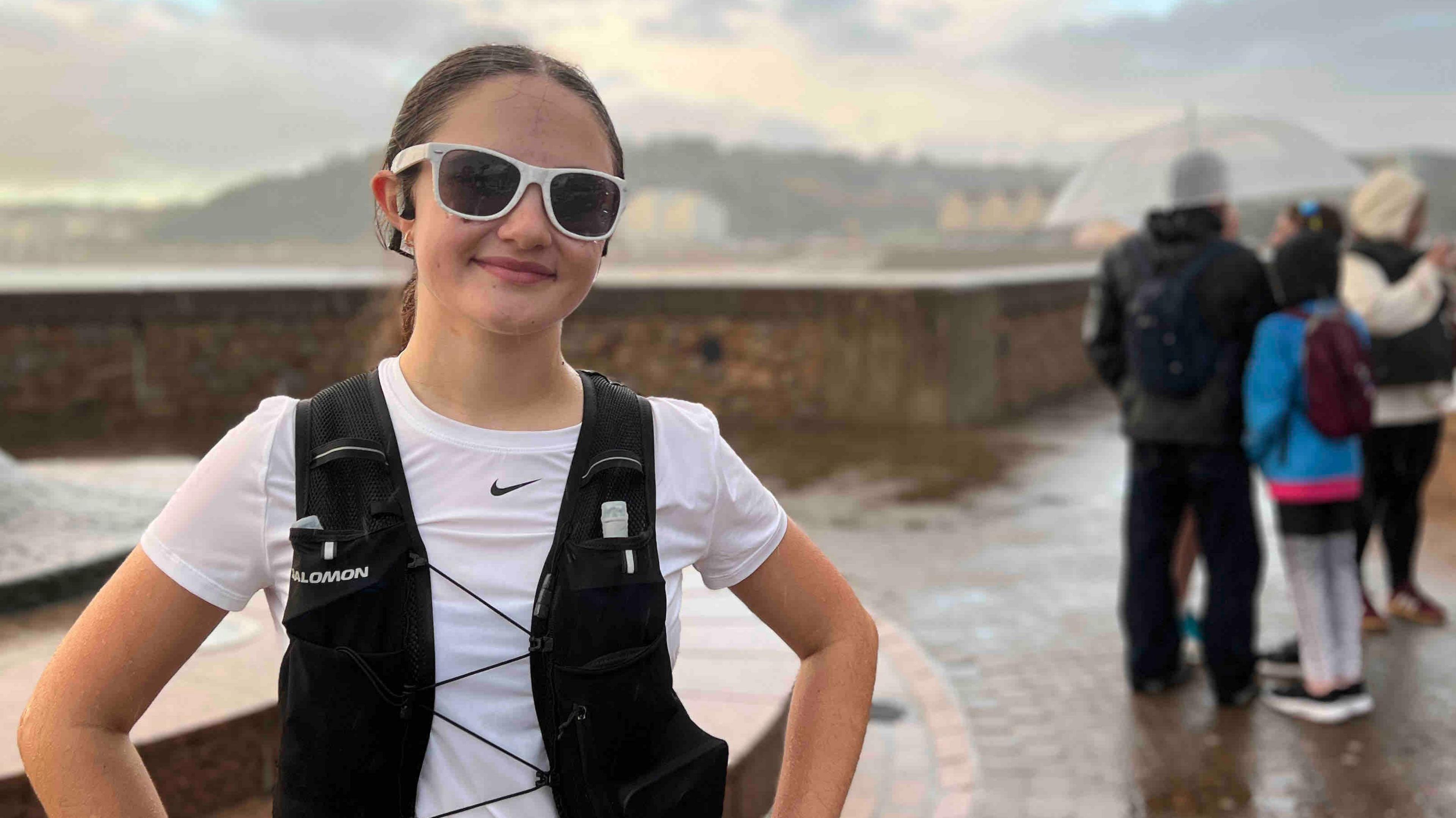 Emily is in a white athletic shirt with a black running vest stands with hands on hips, she is in the foreground. A sports watch is visible on the left wrist. Behind, several people in jackets and hoodies are gathered, some holding umbrellas. The scene overlooks a waterfront area with distant buildings and hills under a cloudy sky, with wet pavement indicating rain.