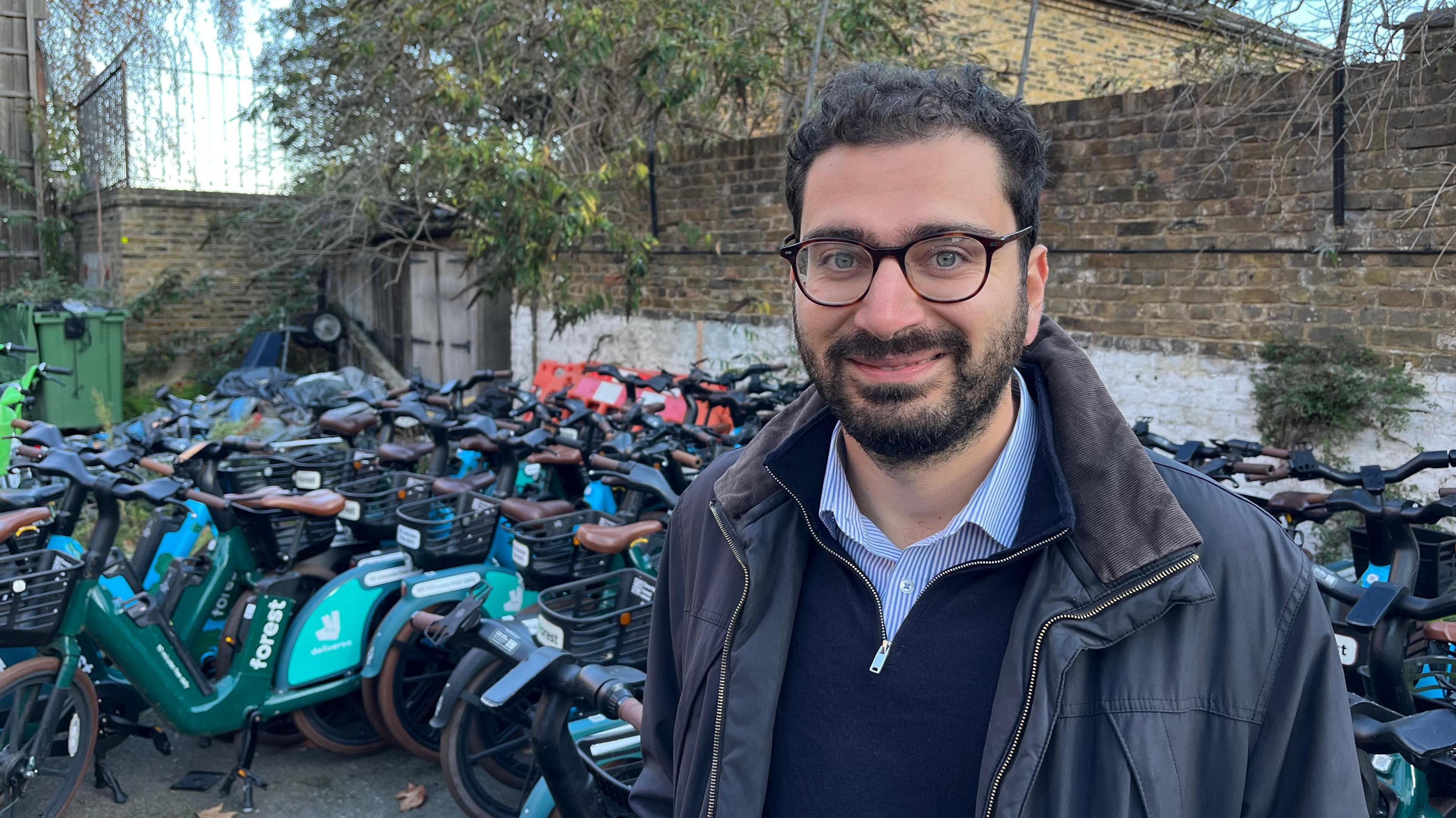 A man with a beard and glasses who is wearing a blue striped shirt beneath a blue top and grey-blue coat is standing in front of lots of e-bikes