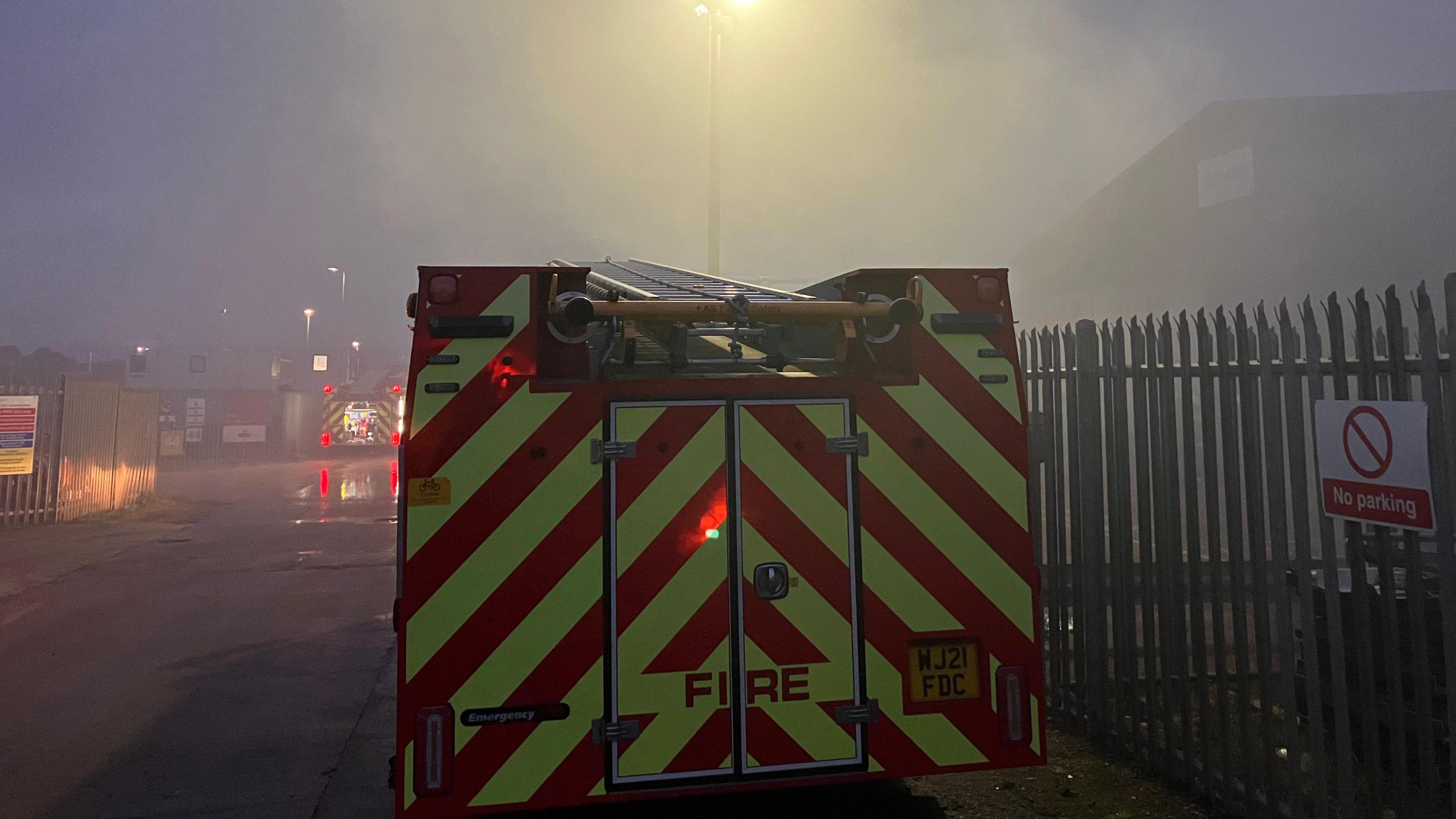 The back of a fire engine parked next to a metal fence which has a sign saying 'no parking on it' with lots of smoke in front of it and the back of a second fire engine visible in the distance