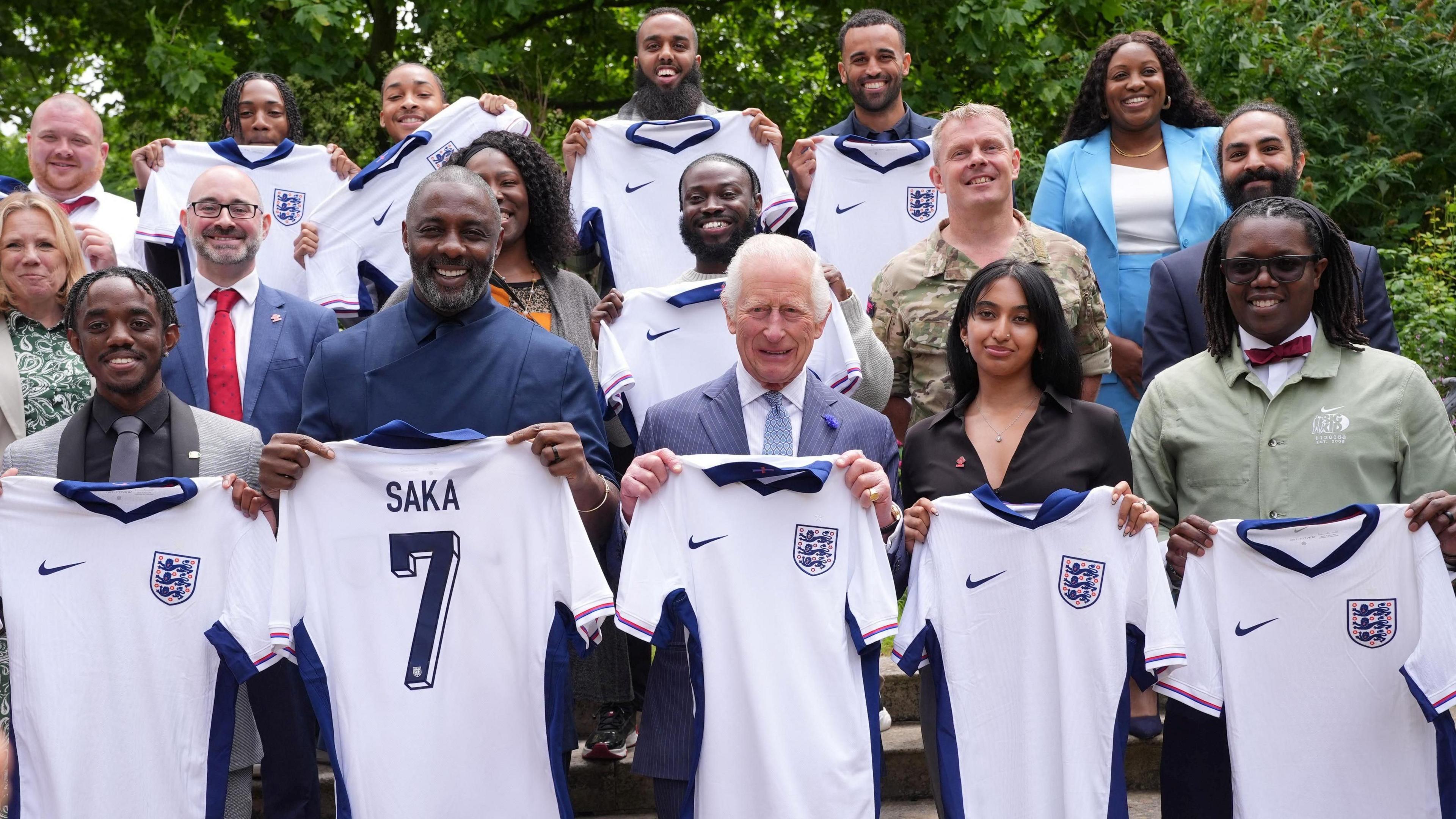 King Charles, Idris Elba, and a group of people stand holding up England football shirts