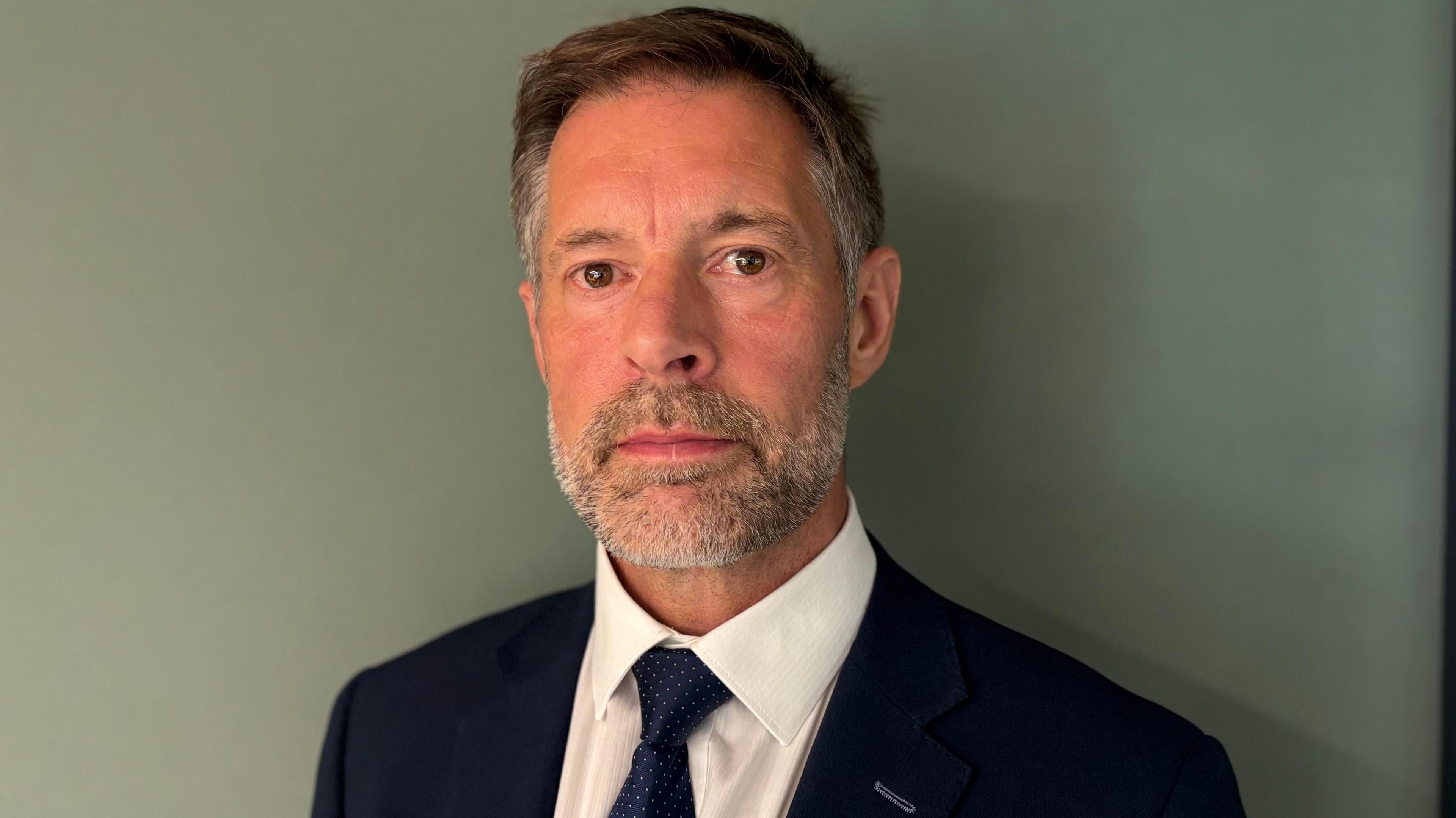 A man in a dark blue suit with white shirt and blue spotted tie. He is standing against a grey wall. He has grey hair and a grey beard.
