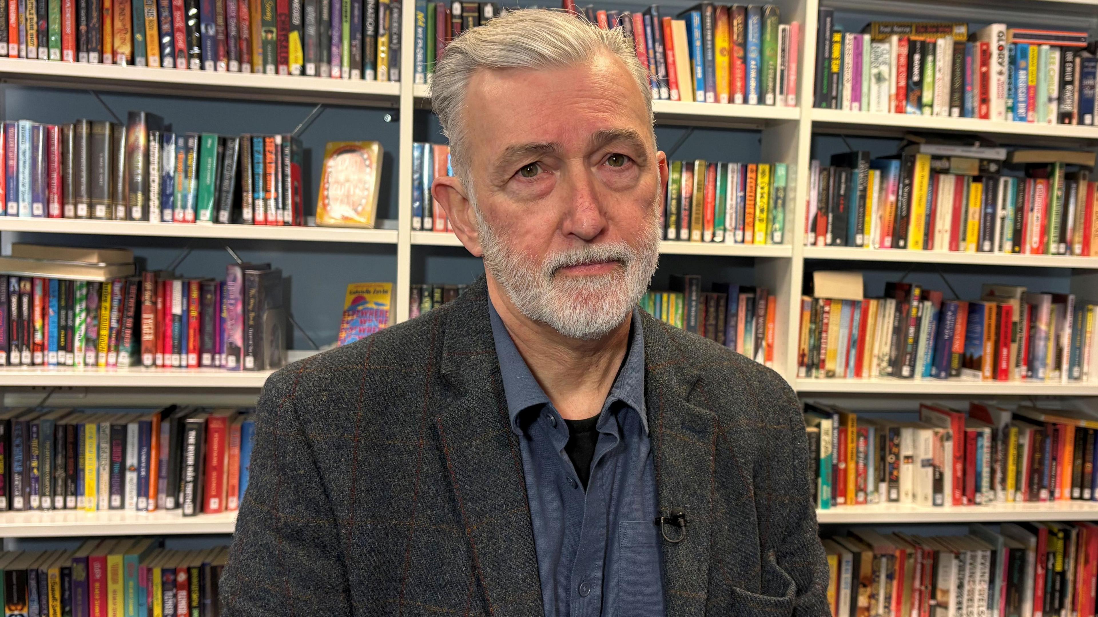 Rod McCready in a school library with books on shelves behind him. He has grey hair and a grey beard. He is wearing a grey wool suit and a blue shirt