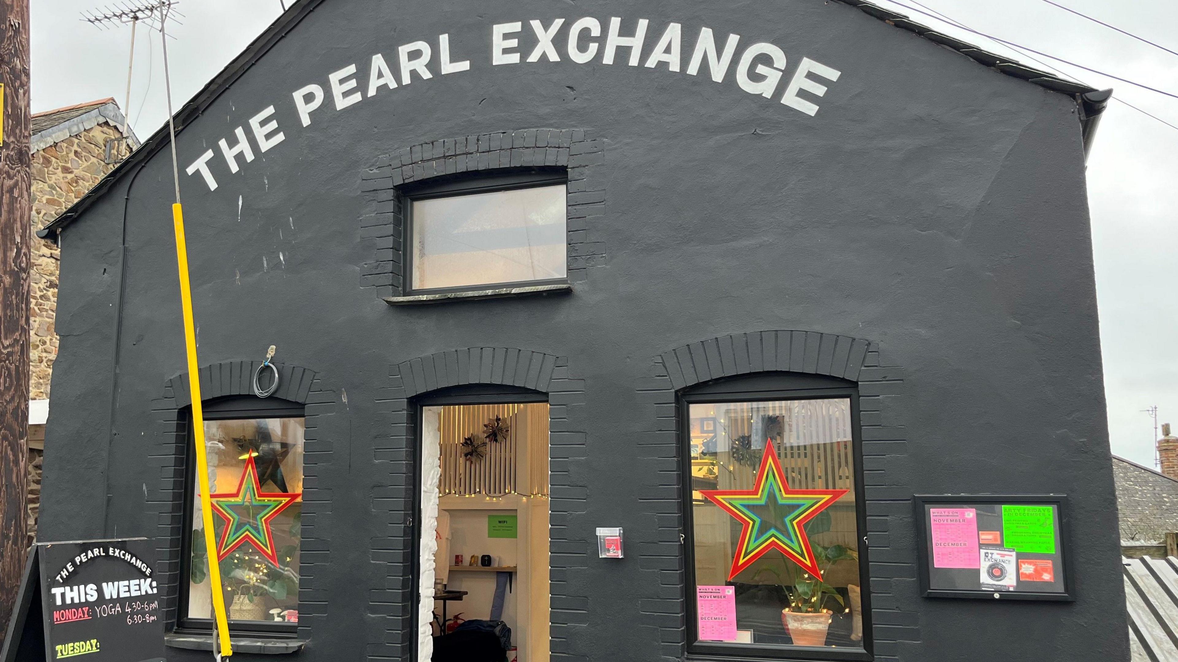 A building with a sloped pointed roof painted black with letters in white at the top spelling out The Pearl Exchange. There are three windows, two have large multi-coloured stars in them, There's a noticeboard with a pink and green posters on the wall and the door is open.