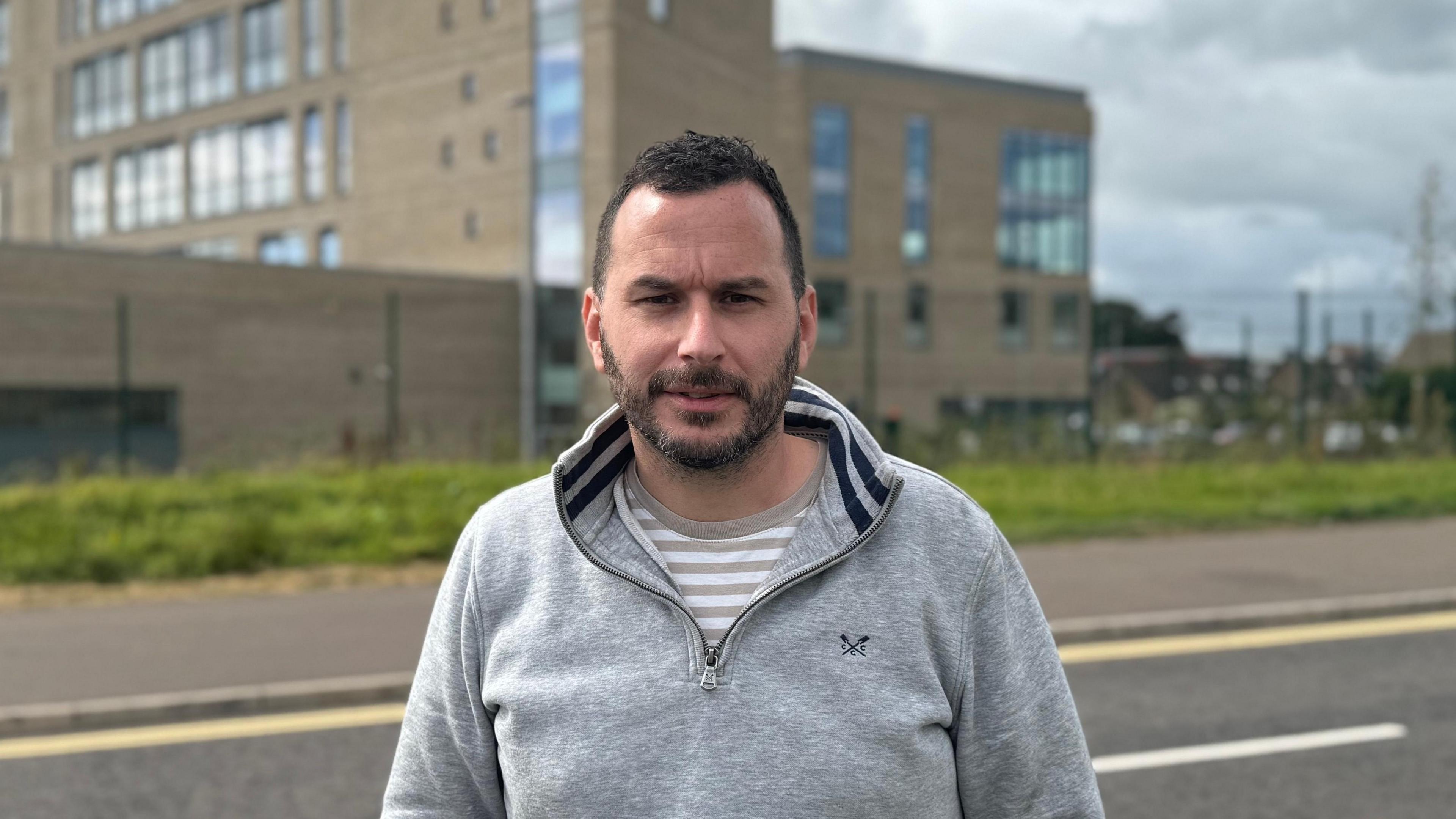 Timothy Gaston is wearing a grey quarter-zip sweatshirt with an upturned collar and a striped white and beige t-shirt underneath it. He has short dark hair and a trimmed beard. He is standing on the roadside staring into the camera. The brown college building is behind him in the distance.