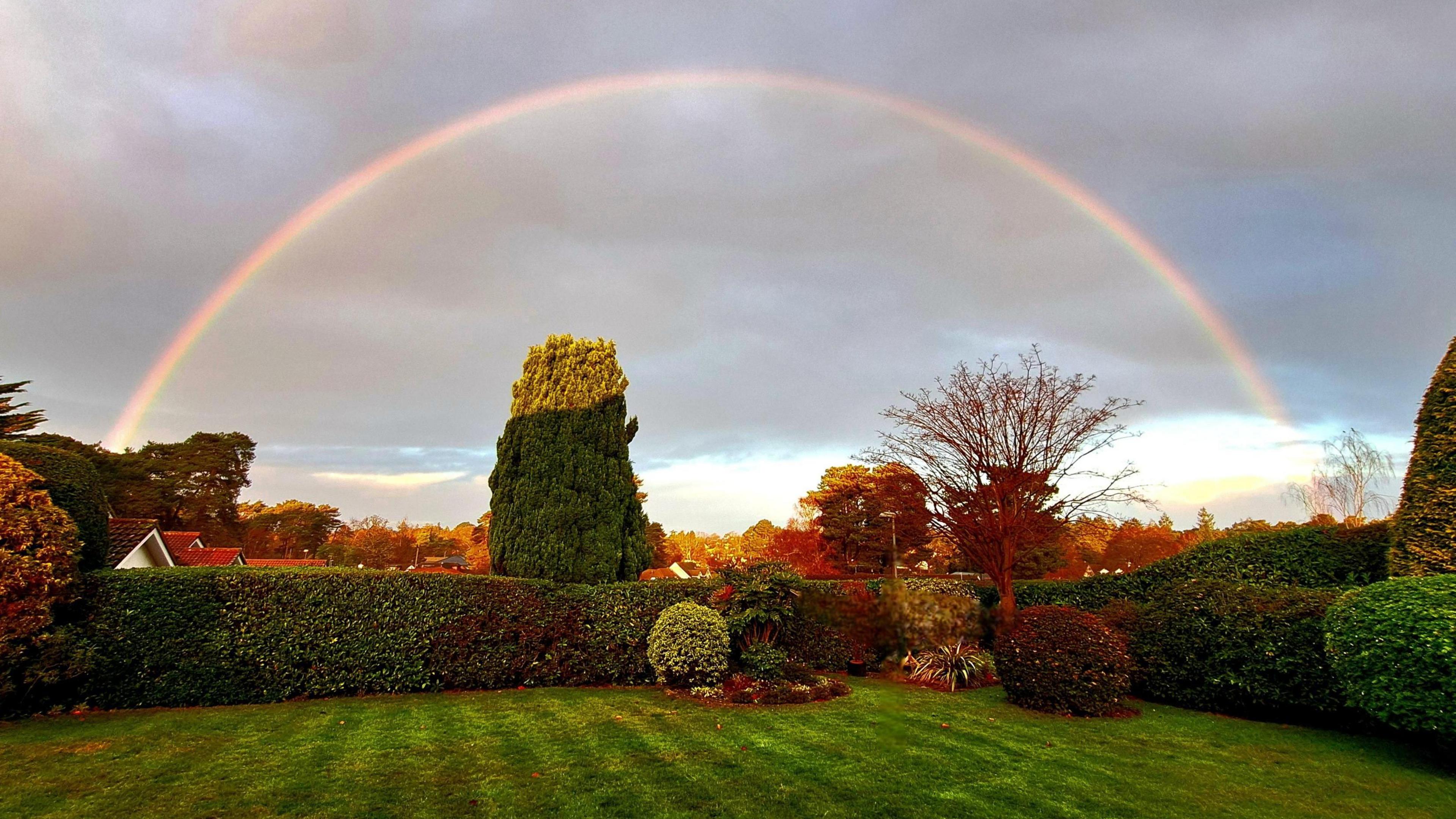 A full rainbow stands out against a grey sky. Underneath it there is a back garden with a green lawn that is surrounded by hedges on all sides. In the distance a few houses can be seen and some trees of varying autumnal colours.