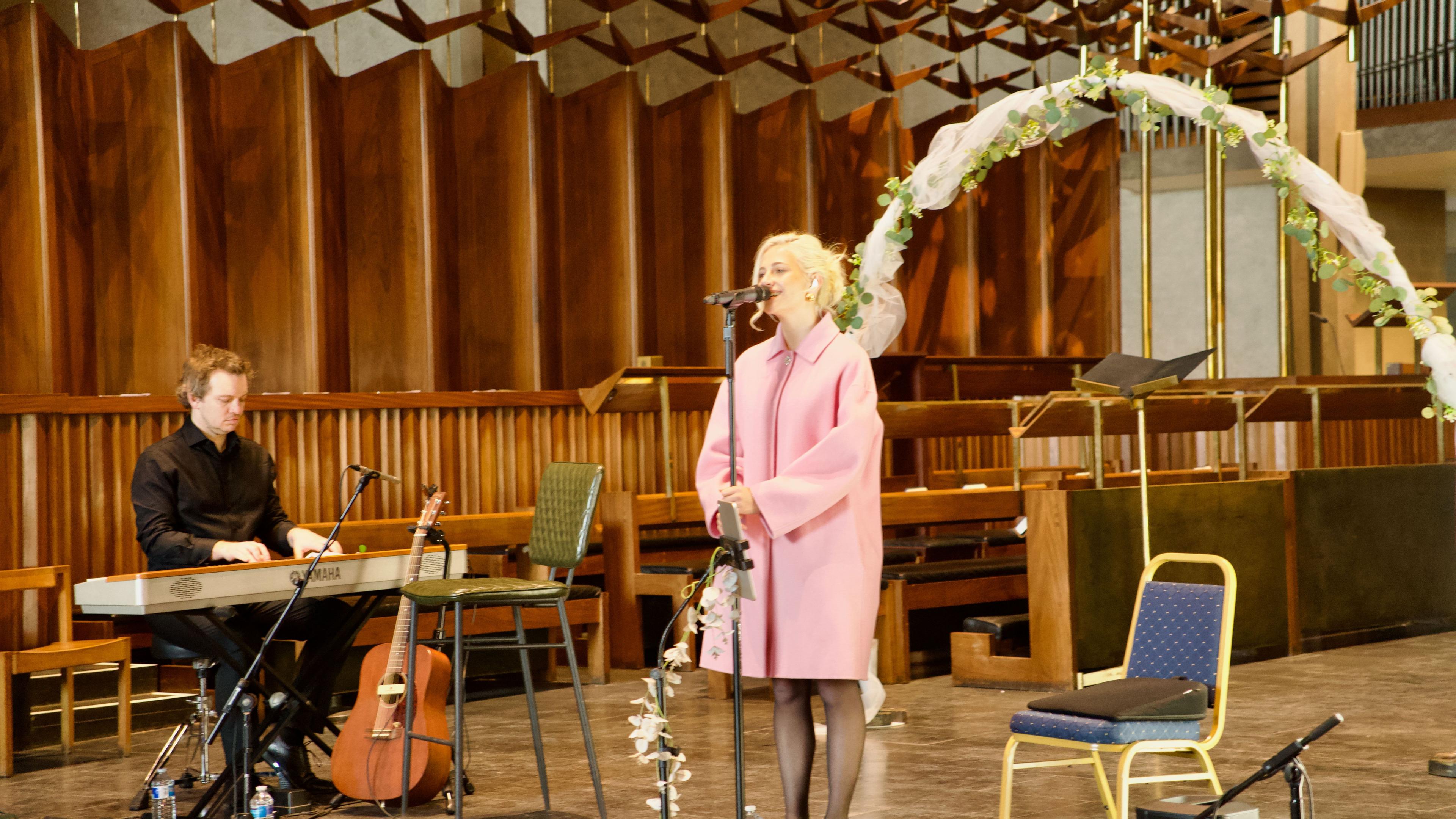 Singer in a pink coat performs at Coventry Cathedral. To her right is a keyboard player in a black shirt