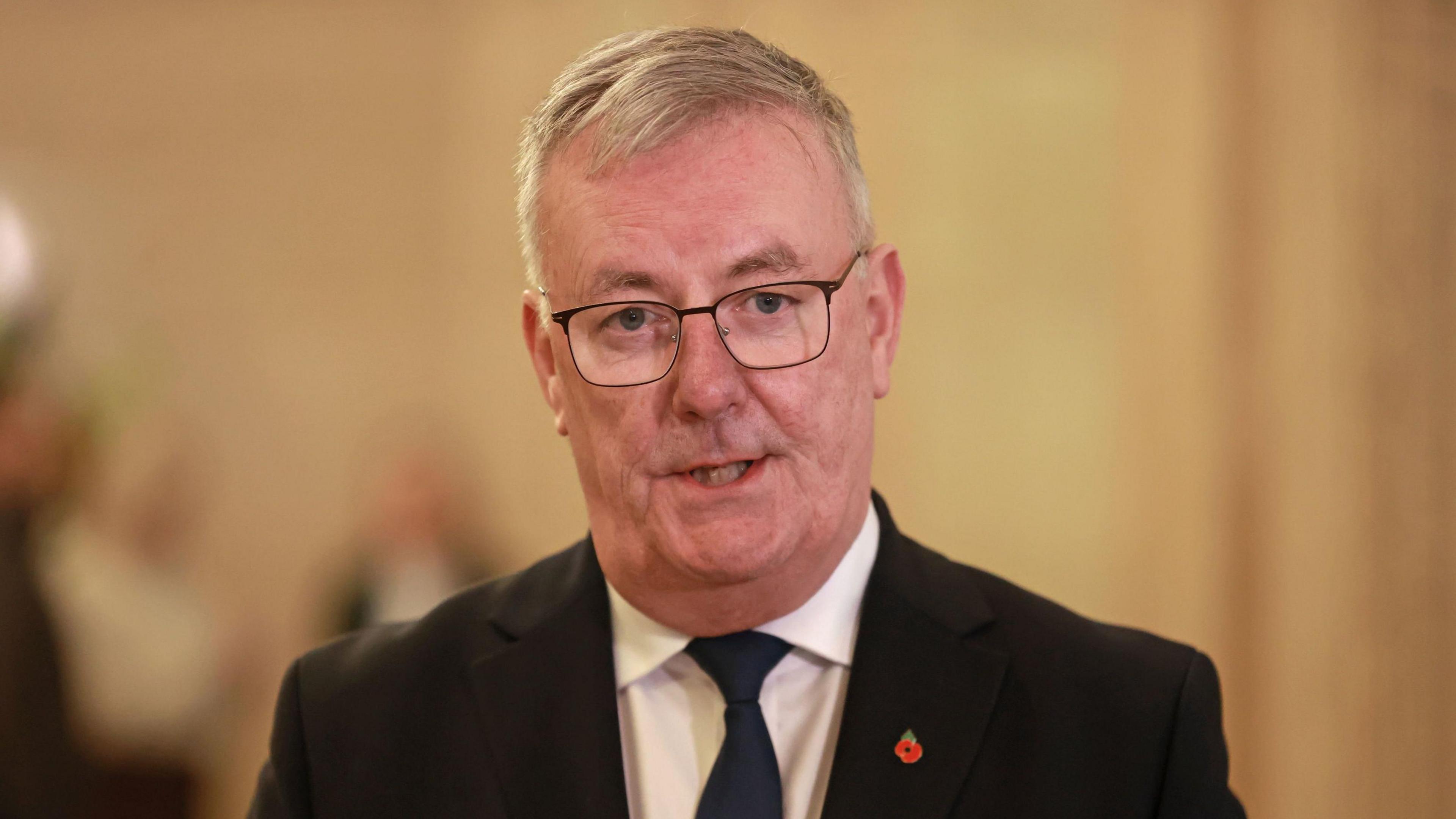 Mike Nesbitt stands in Stormont. He has grey hair and glasses, he is dressed in a black suit with a white shirt and blue tie. Behind him there are white walls.