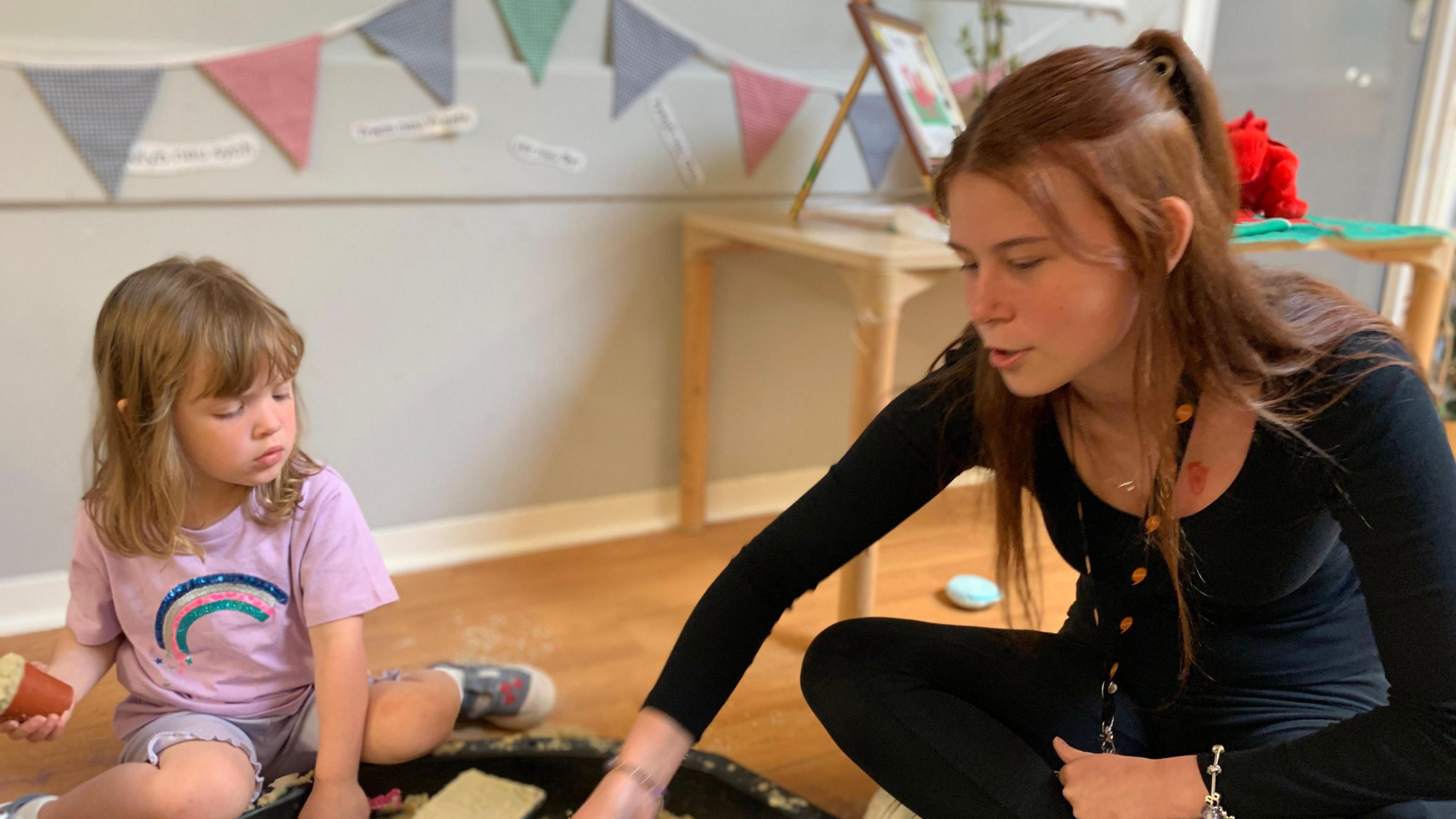 A woman and a child playing in a room. The woman is to the right of the image and is wearing a black top and black leggings, she has shoulder-length red hair and has a black lanyard with orange speech bubbles on it. The child is on the left and has a purple t-shirt on with a rainbow on it and purple shorts. She has blonde shoulder-length hair.