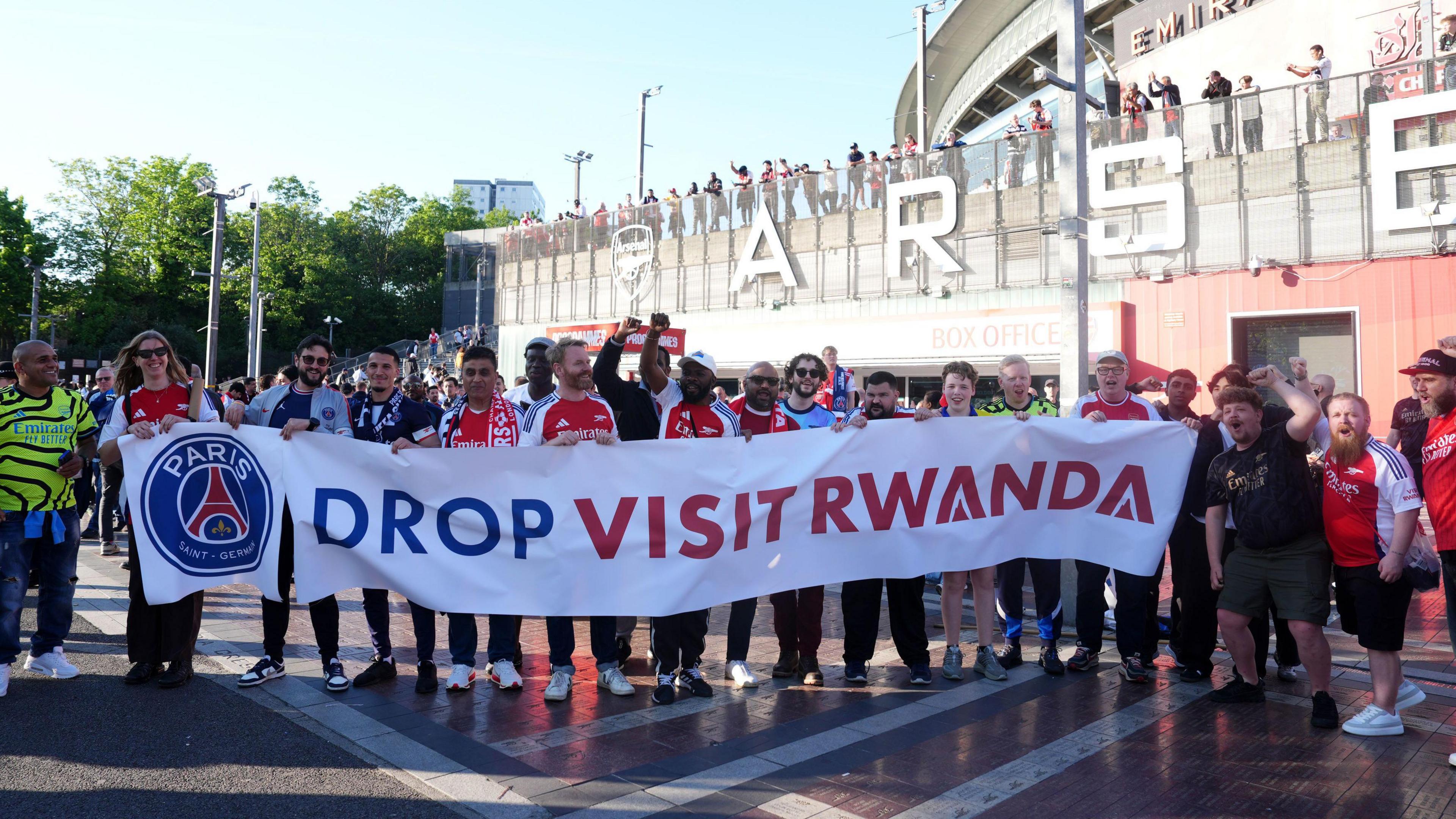 Arsenal and PSG fans stand behind a banner reading 'Drop Visit Rwanda' outside the Emirates Stadium